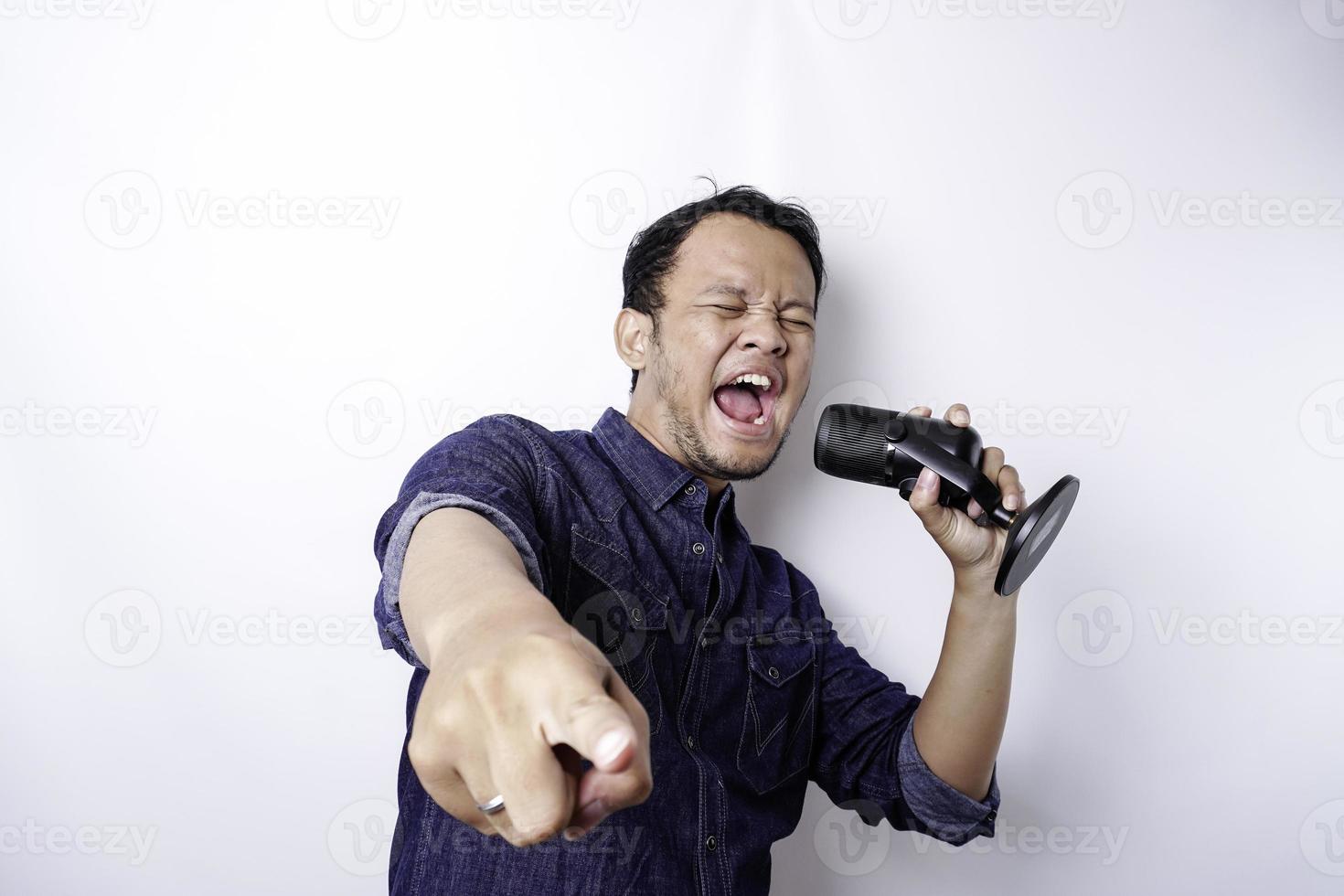 Portrait of carefree Asian man, having fun karaoke, singing in microphone while standing over white background photo