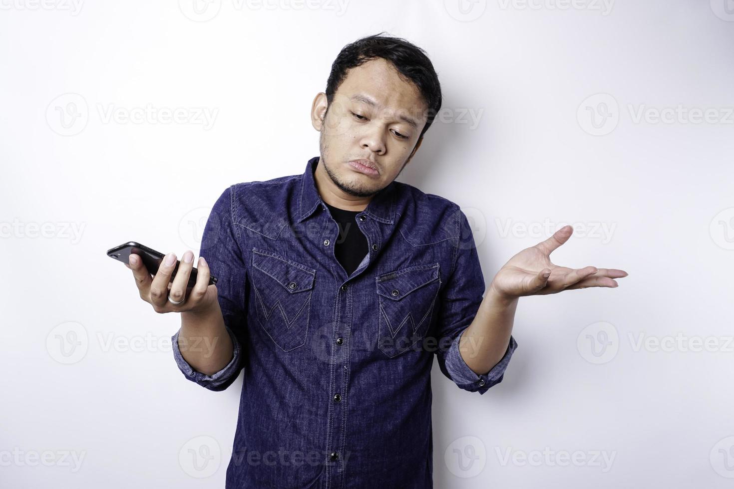 A thoughtful young Asian man is wearing blue shirt holding his phone and looks confused, isolated by white background photo