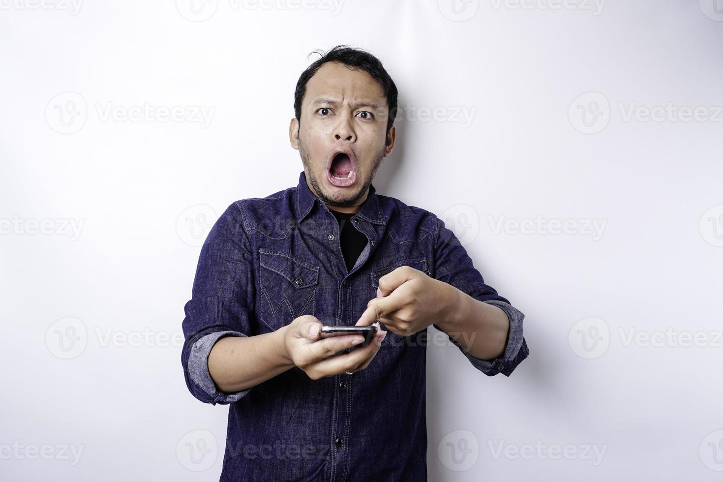 hombre asiático conmocionado con camisa azul y sosteniendo su teléfono, aislado de fondo blanco foto