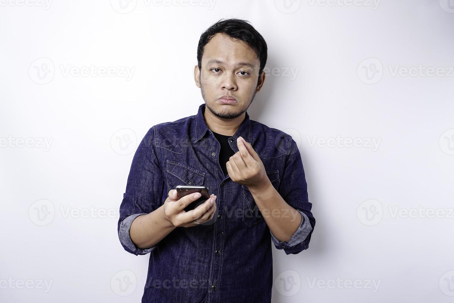 A thoughtful young man dressed in blue shirt while looking aside holding his phone, isolated by white background photo
