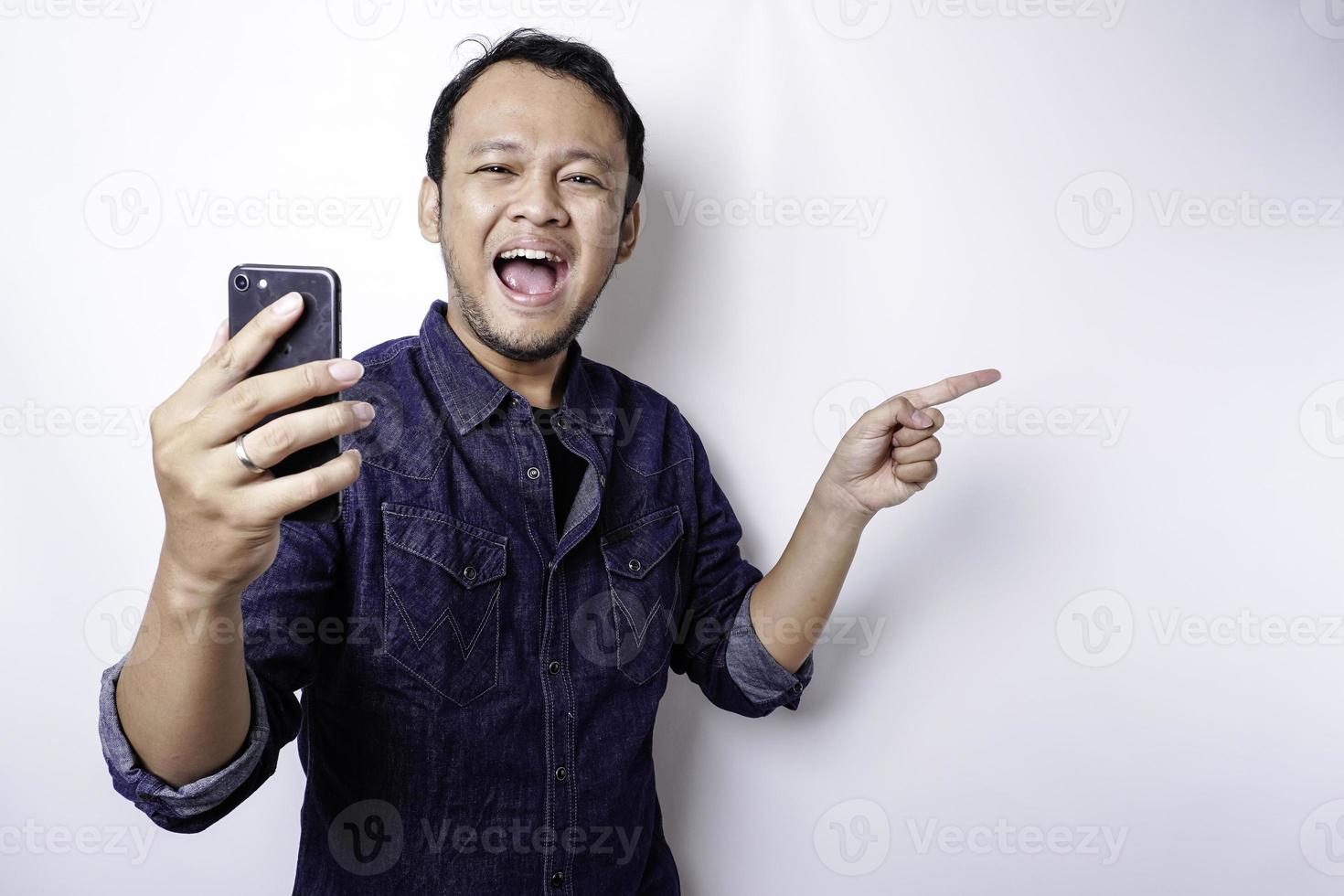 Excited Asian man wearing blue shirt pointing at the copy space beside him while holding his phone, isolated by white background photo