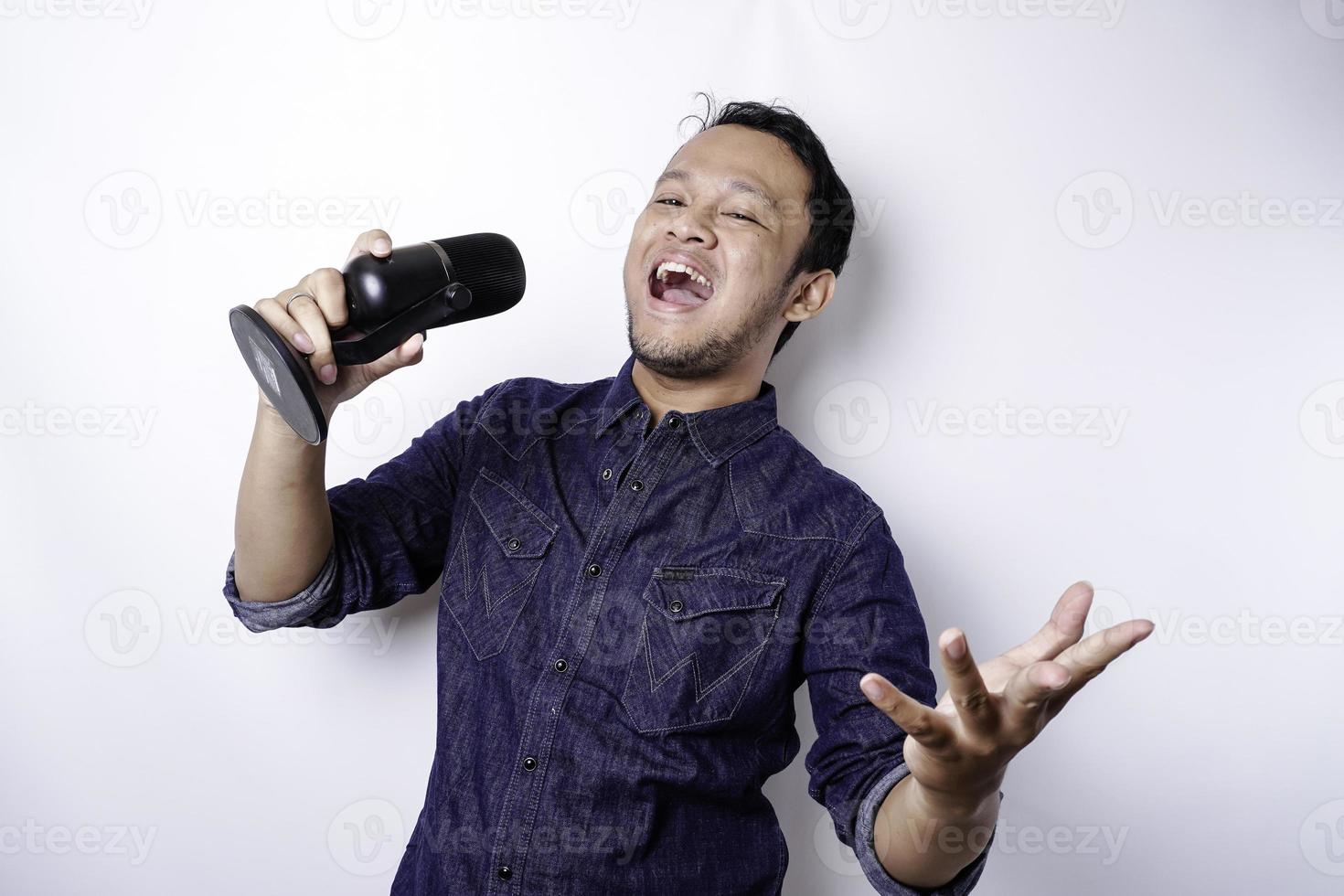 Portrait of carefree Asian man, having fun karaoke, singing in microphone while standing over white background photo