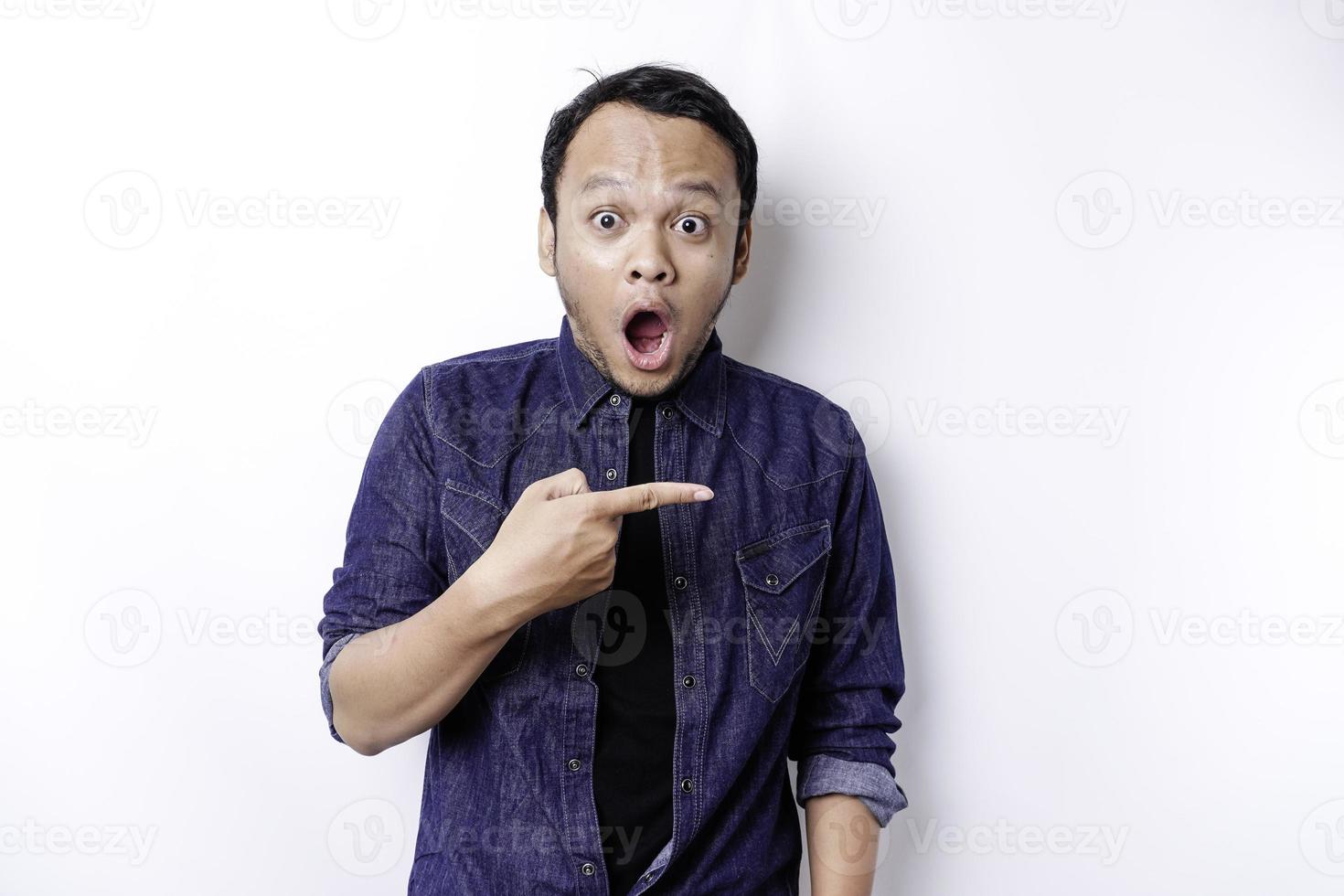 Shocked Asian man wearing blue shirt pointing at the copy space beside him, isolated by white background photo