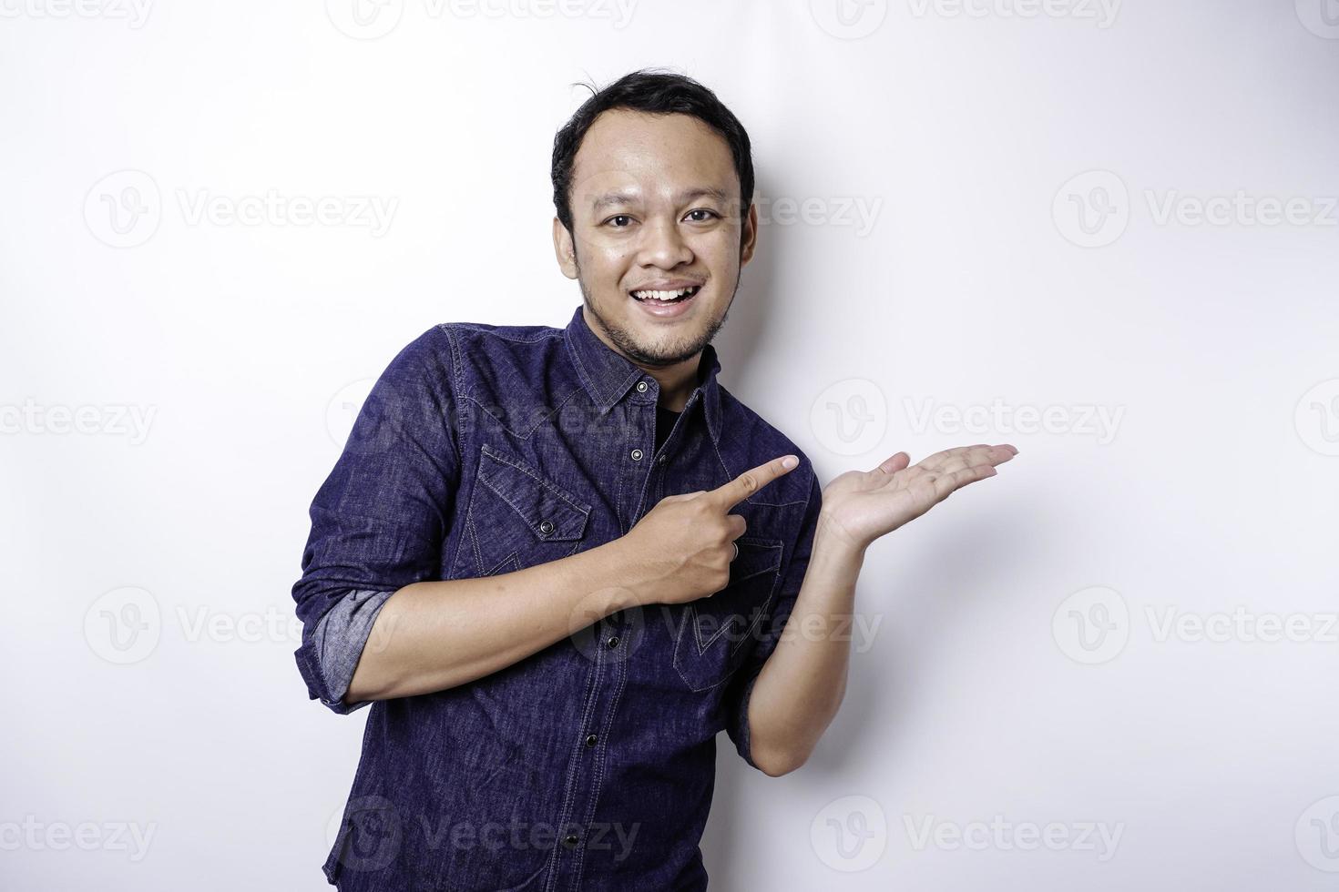 Excited Asian man wearing blue shirt pointing at the copy space beside him, isolated by white background photo