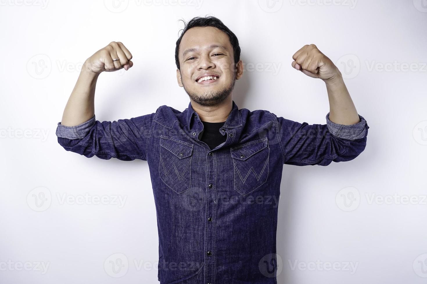 hombre asiático emocionado con una camisa azul que muestra un gesto fuerte levantando los brazos y los músculos sonriendo con orgullo foto