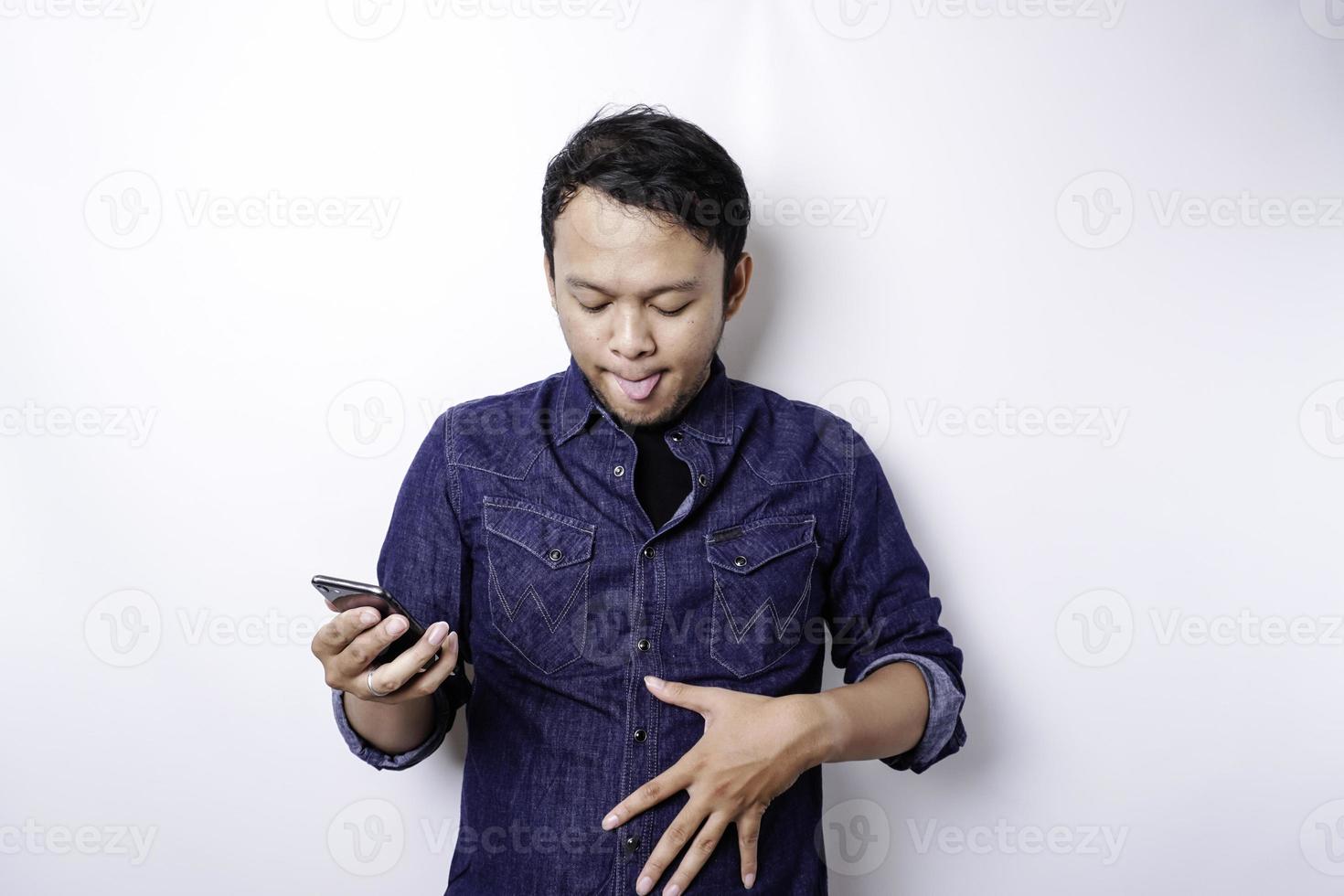 un hombre asiático pensativo tiene hambre y se toca la barriga mientras sostiene su teléfono pensando qué pedir, aislado de fondo blanco foto