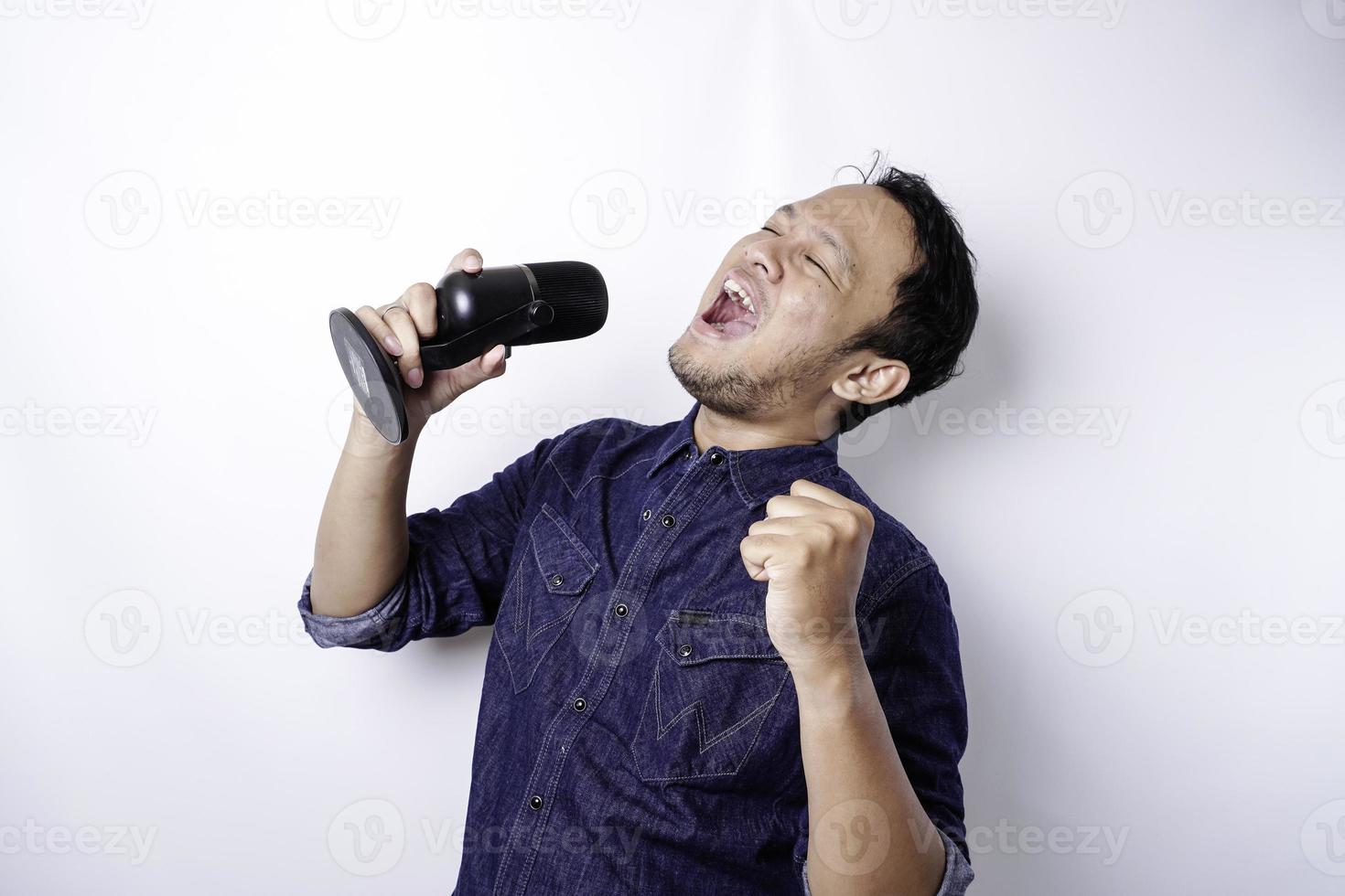 Portrait of carefree Asian man, having fun karaoke, singing in microphone while standing over white background photo