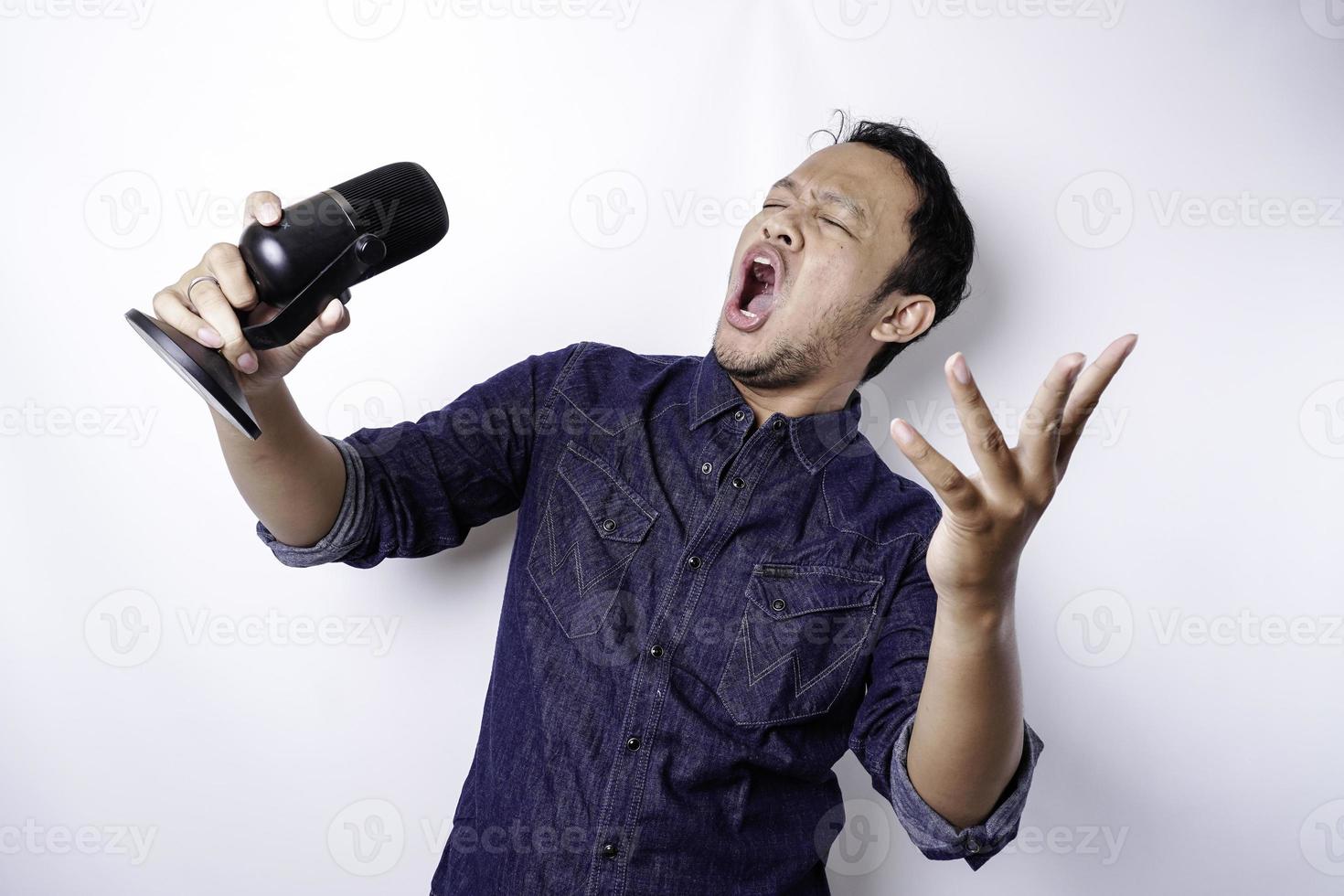 Portrait of carefree Asian man, having fun karaoke, singing in microphone while standing over white background photo