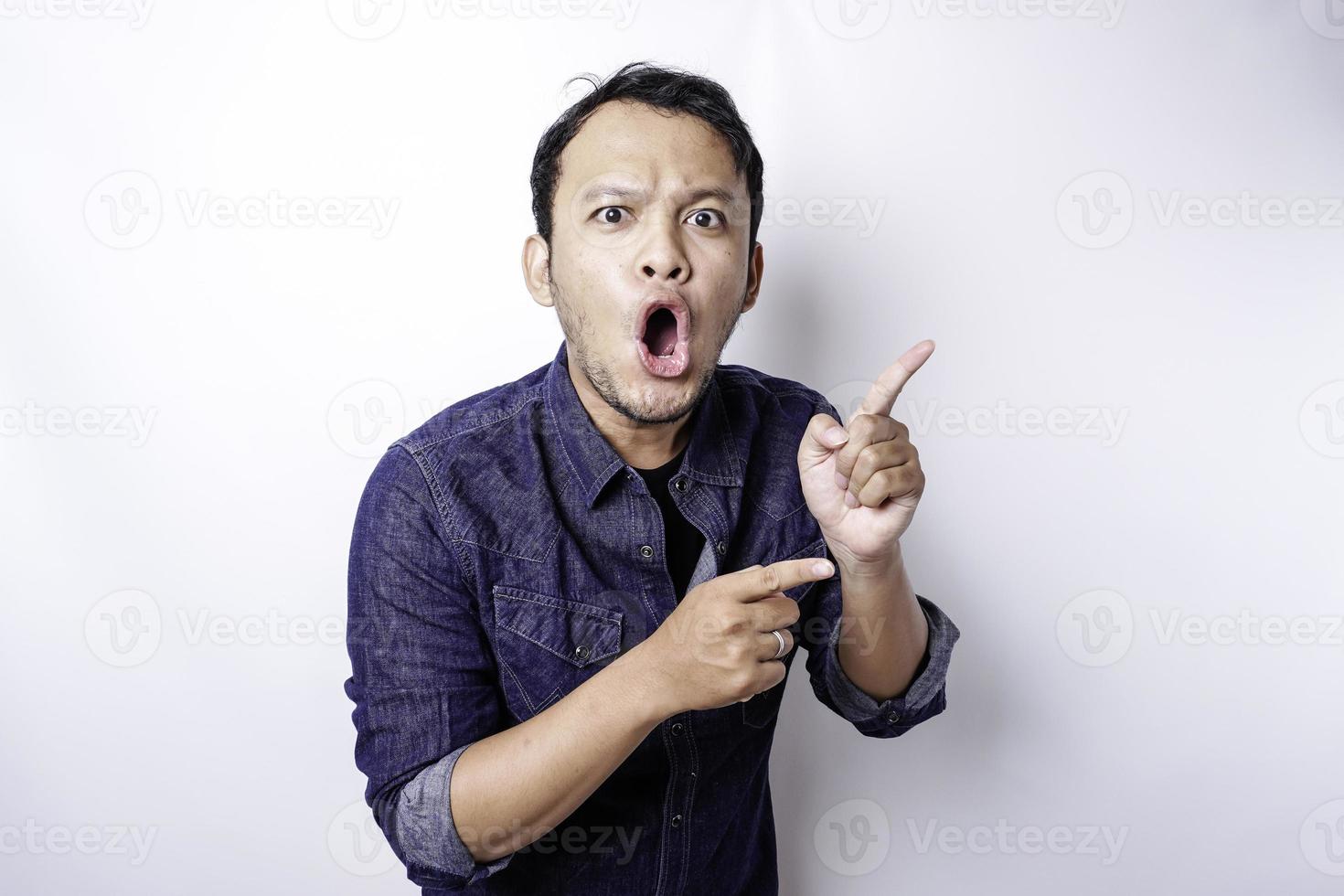 Shocked Asian man wearing blue shirt pointing at the copy space beside him, isolated by white background photo