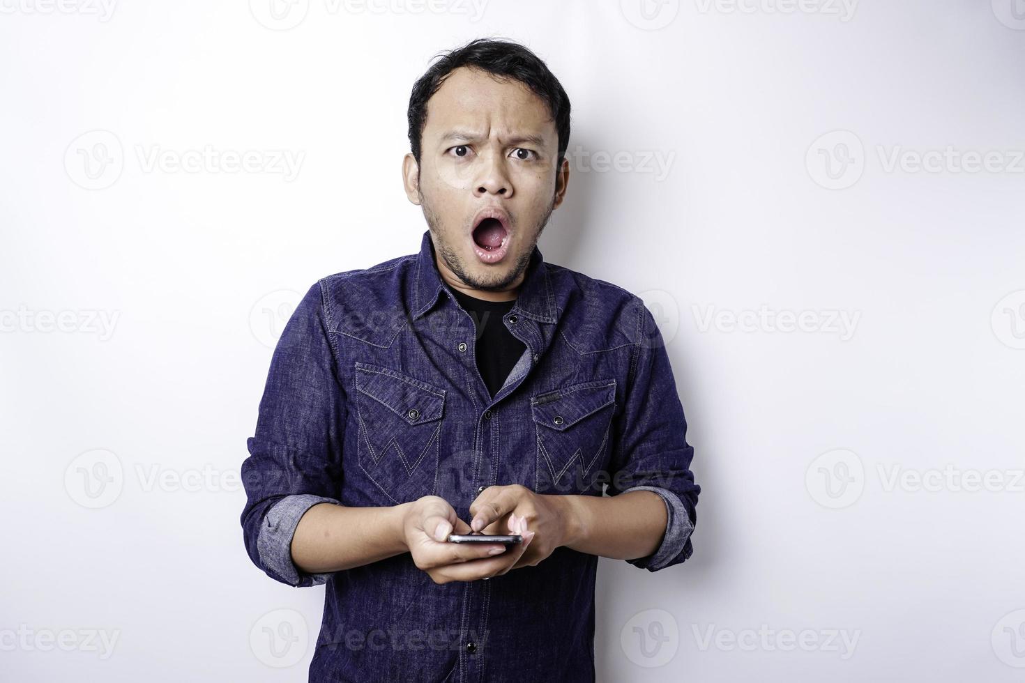 hombre asiático conmocionado con camisa azul y sosteniendo su teléfono, aislado de fondo blanco foto