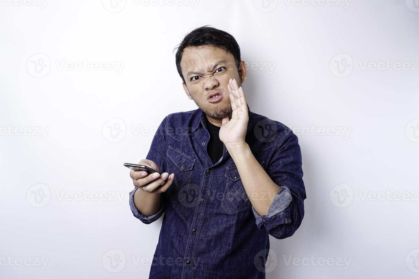 la cara enojada y enojada del hombre asiático con camisa azul sosteniendo su teléfono aislado de fondo blanco. foto