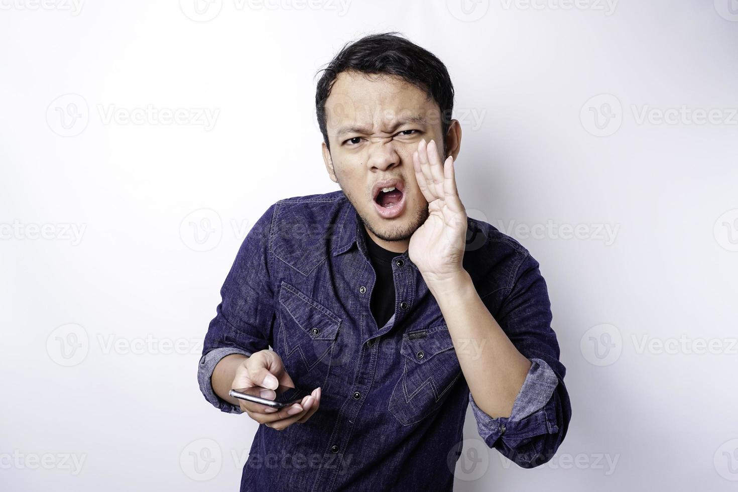 The angry and mad face of Asian man in blue shirt holding his phone isolated white background. photo