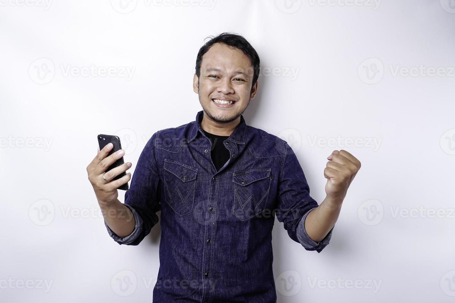 A young Asian man with a happy successful expression wearing blue shirt and holding his phone, isolated by white background photo