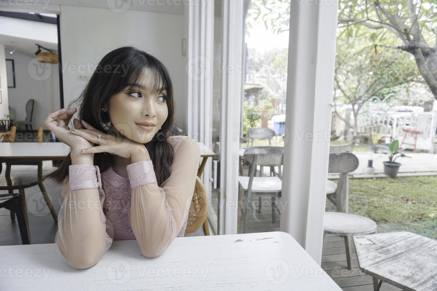 Thoughtful Asian woman keeps hand on chin looks pensively above dressed in pink dress poses thinks about future photo