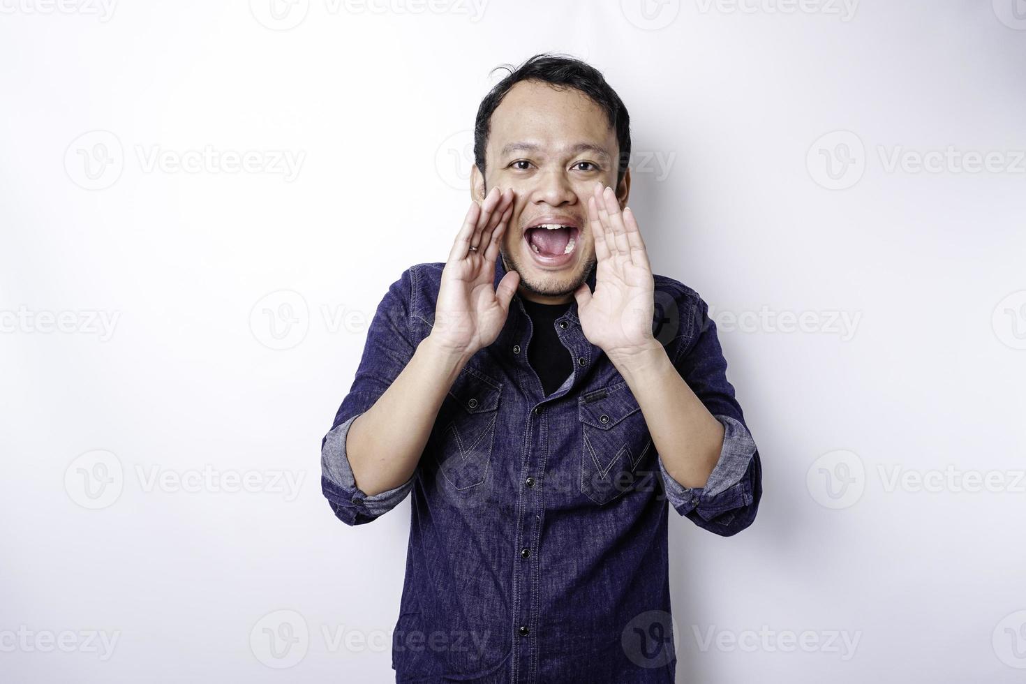 joven apuesto con una camisa azul gritando y gritando fuerte con una mano en la boca. concepto de comunicación foto