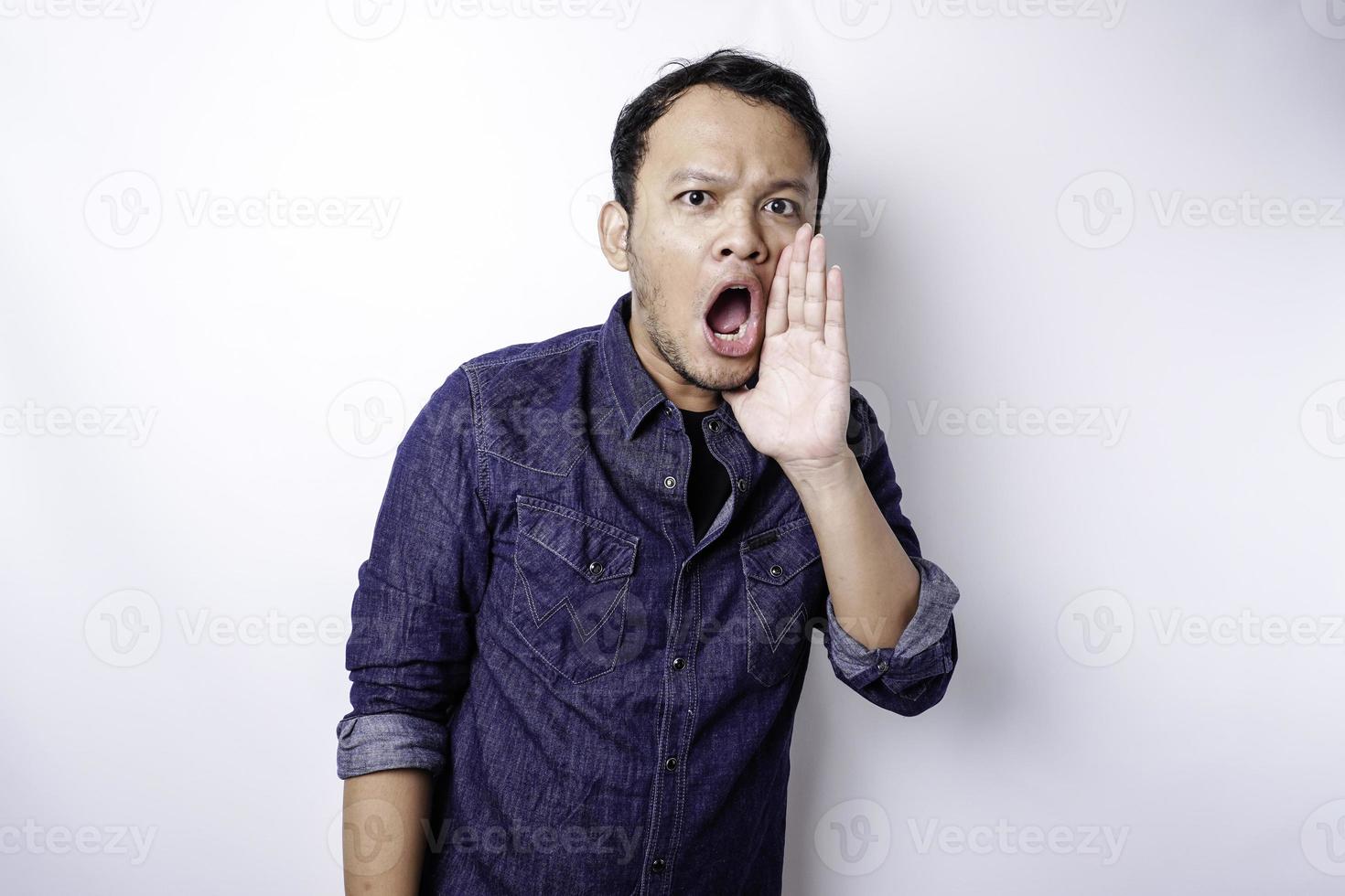 joven apuesto con una camisa azul gritando y gritando fuerte con una mano en la boca. concepto de comunicación foto