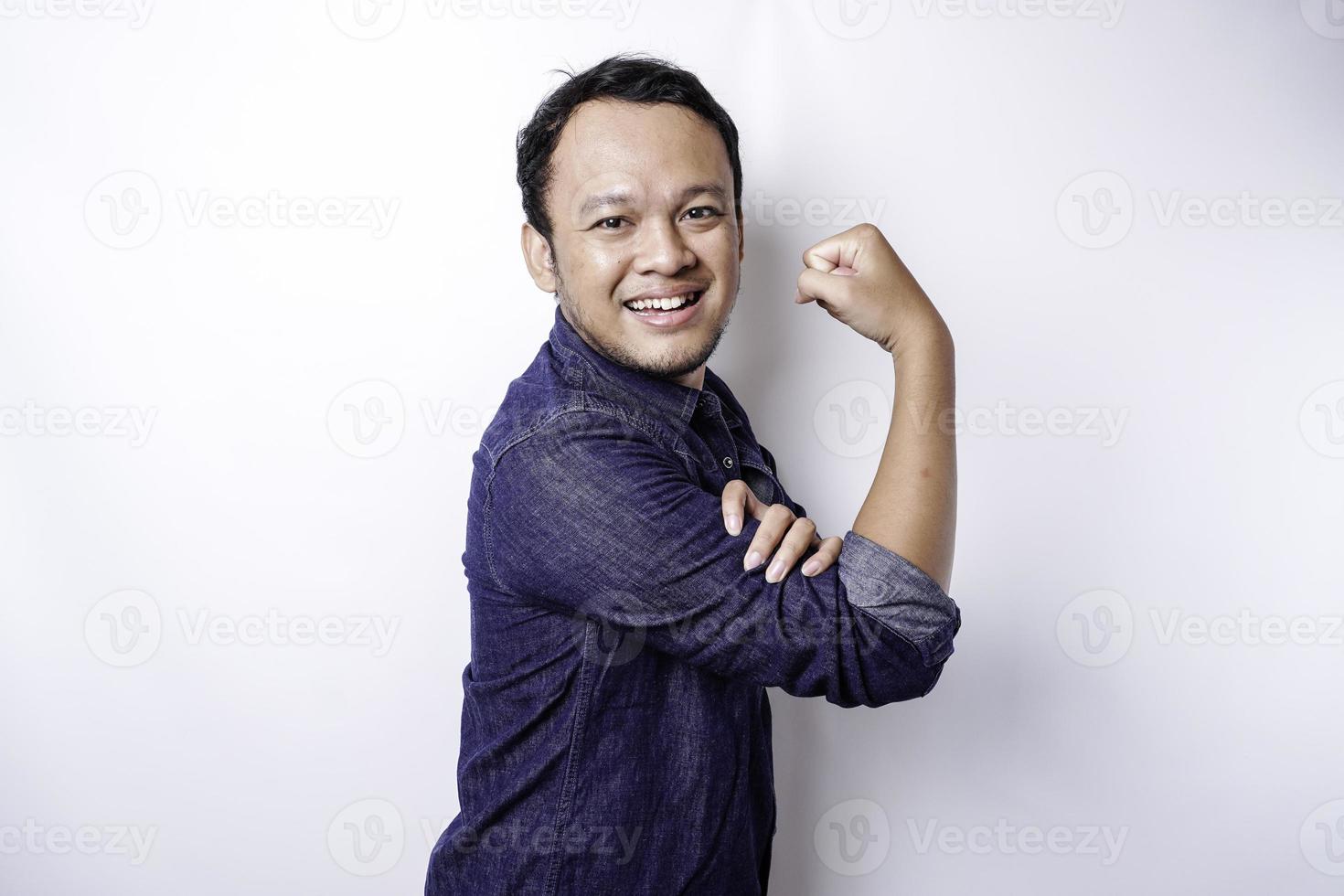 Excited Asian man wearing a blue shirt showing strong gesture by lifting his arms and muscles smiling proudly photo