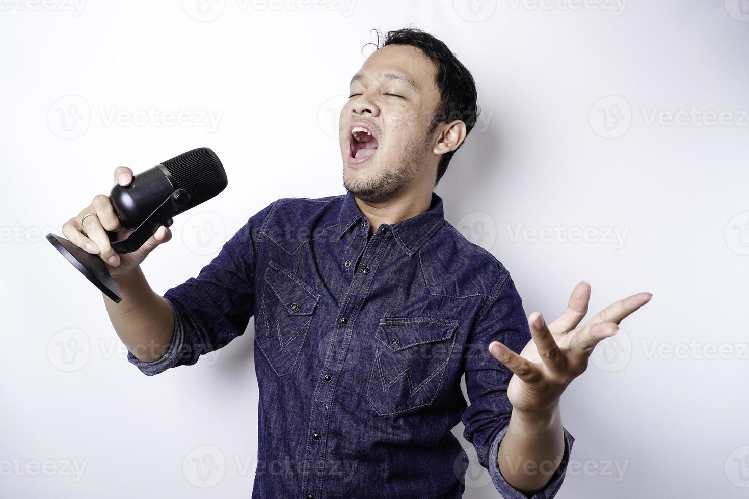 Portrait of carefree Asian man, having fun karaoke, singing in microphone while standing over white background photo