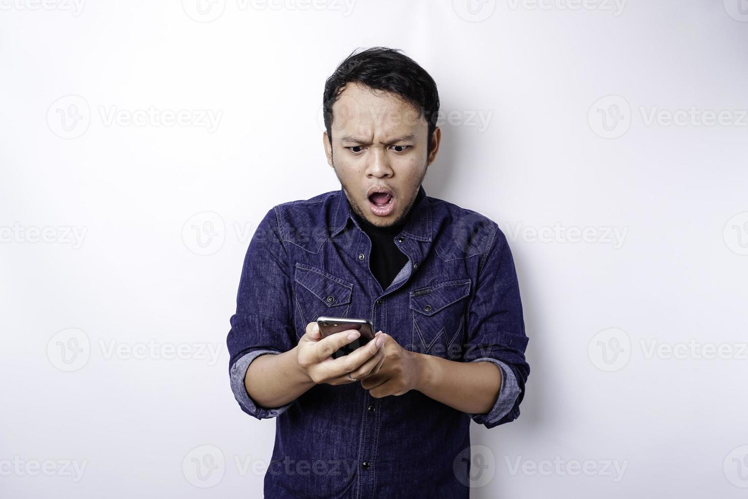 Shocked Asian man wearing blue shirt and holding his phone, isolated by white background photo