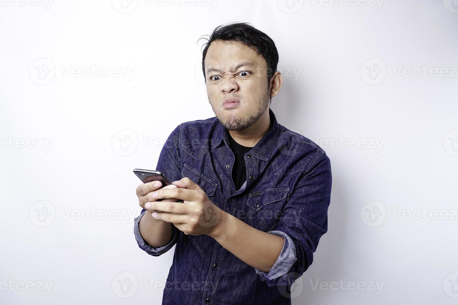 The angry and mad face of Asian man in blue shirt holding his phone isolated white background. photo