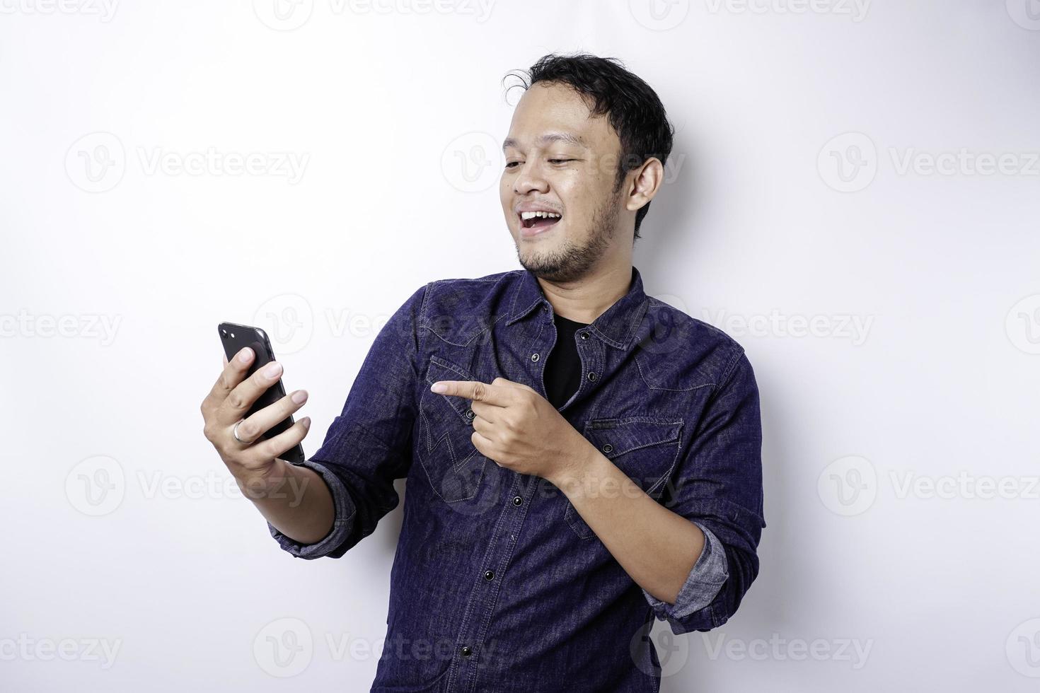 Excited Asian man wearing blue shirt smiling while holding his phone, isolated by white background photo