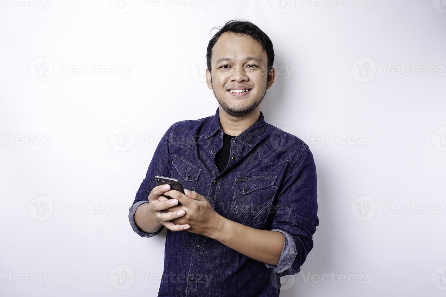 Excited Asian man wearing blue shirt smiling while holding his phone, isolated by white background photo