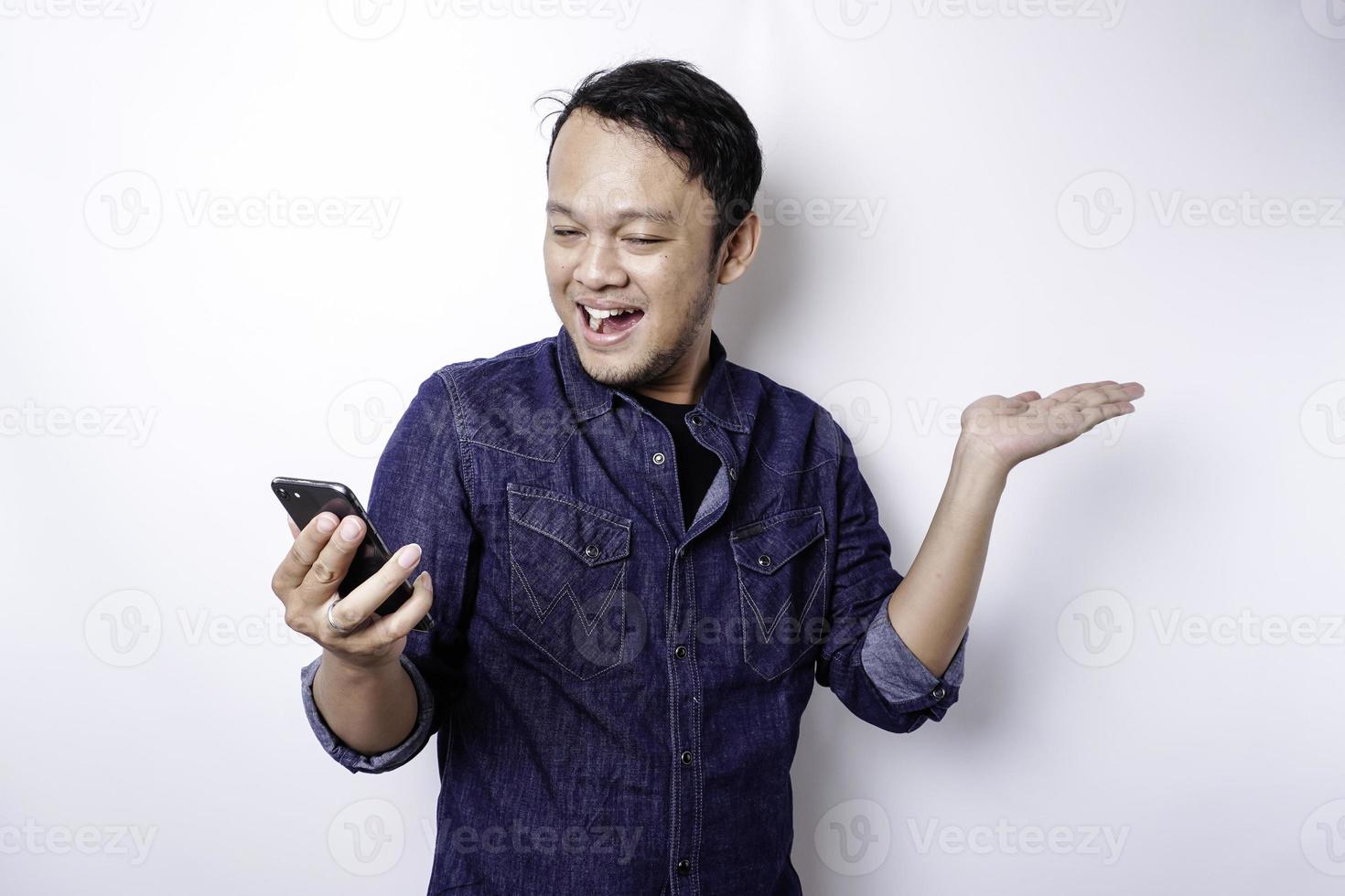 Excited Asian man wearing blue shirt pointing at the copy space beside him while holding his phone, isolated by white background photo
