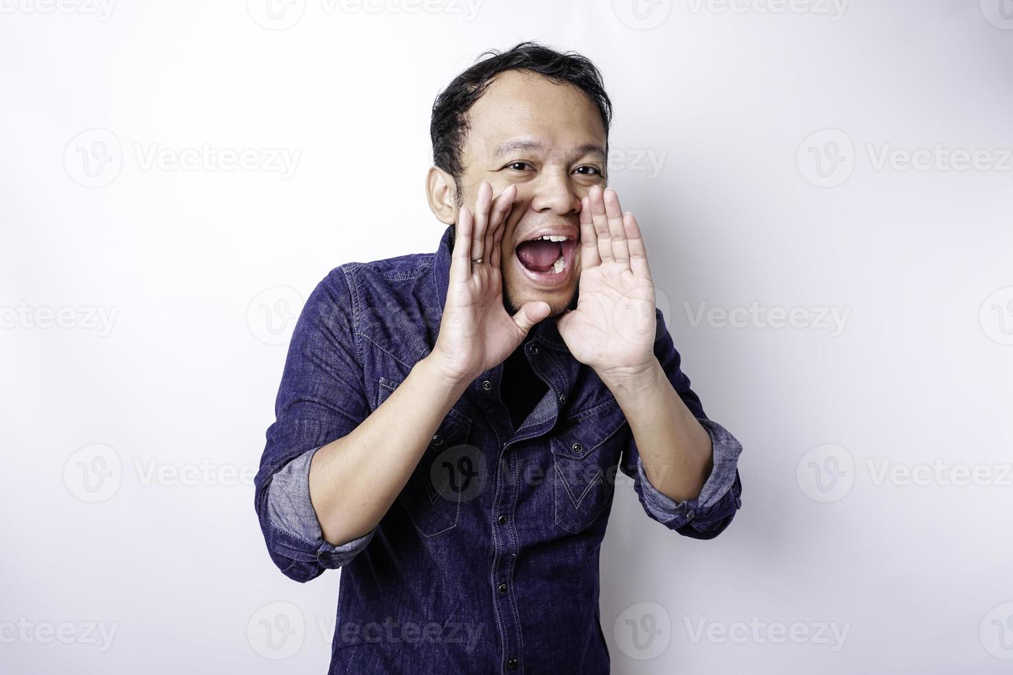 joven apuesto con una camisa azul gritando y gritando fuerte con una mano en la boca. concepto de comunicación foto