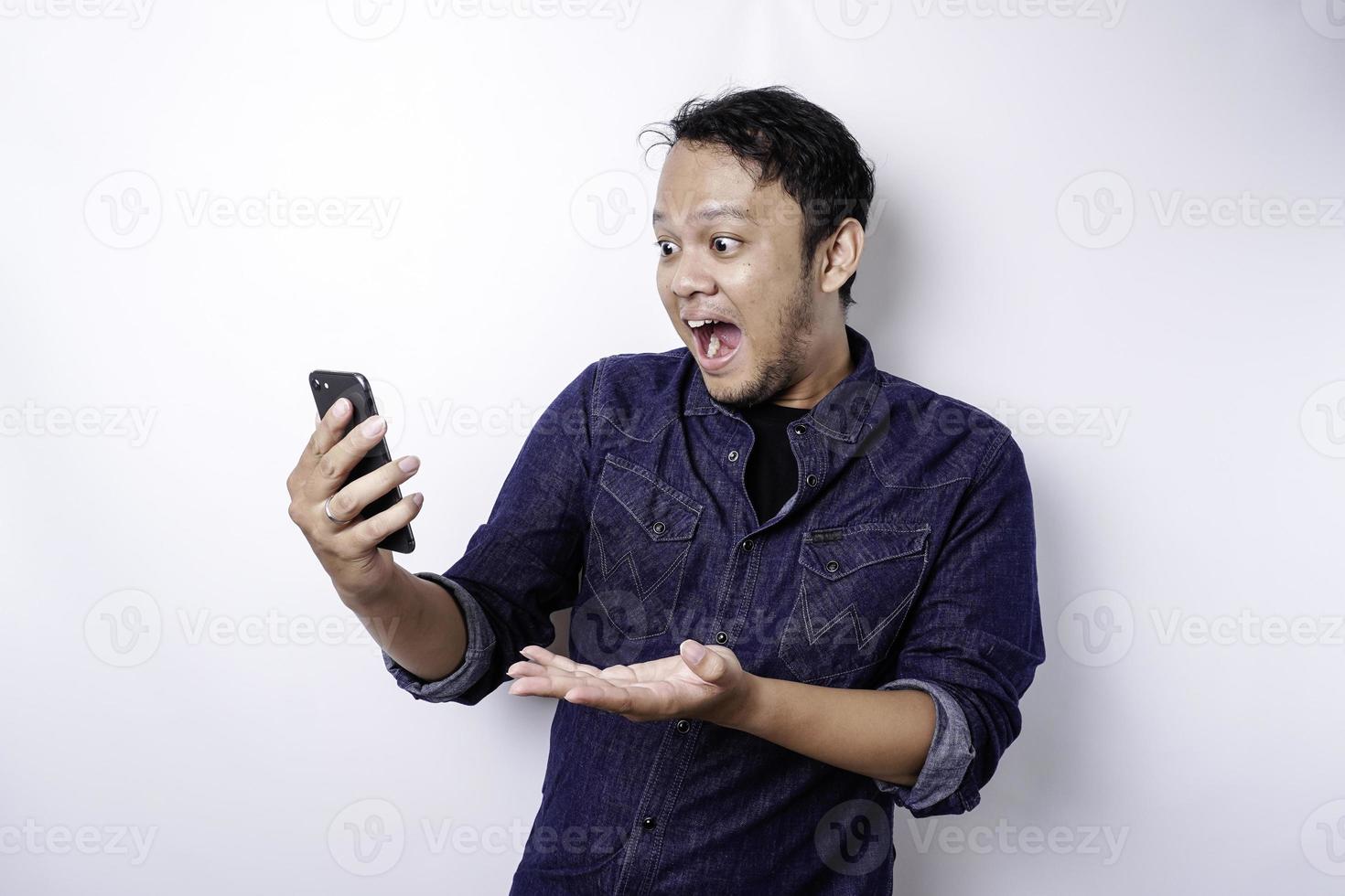 hombre asiático conmocionado con camisa azul y sosteniendo su teléfono, aislado de fondo blanco foto