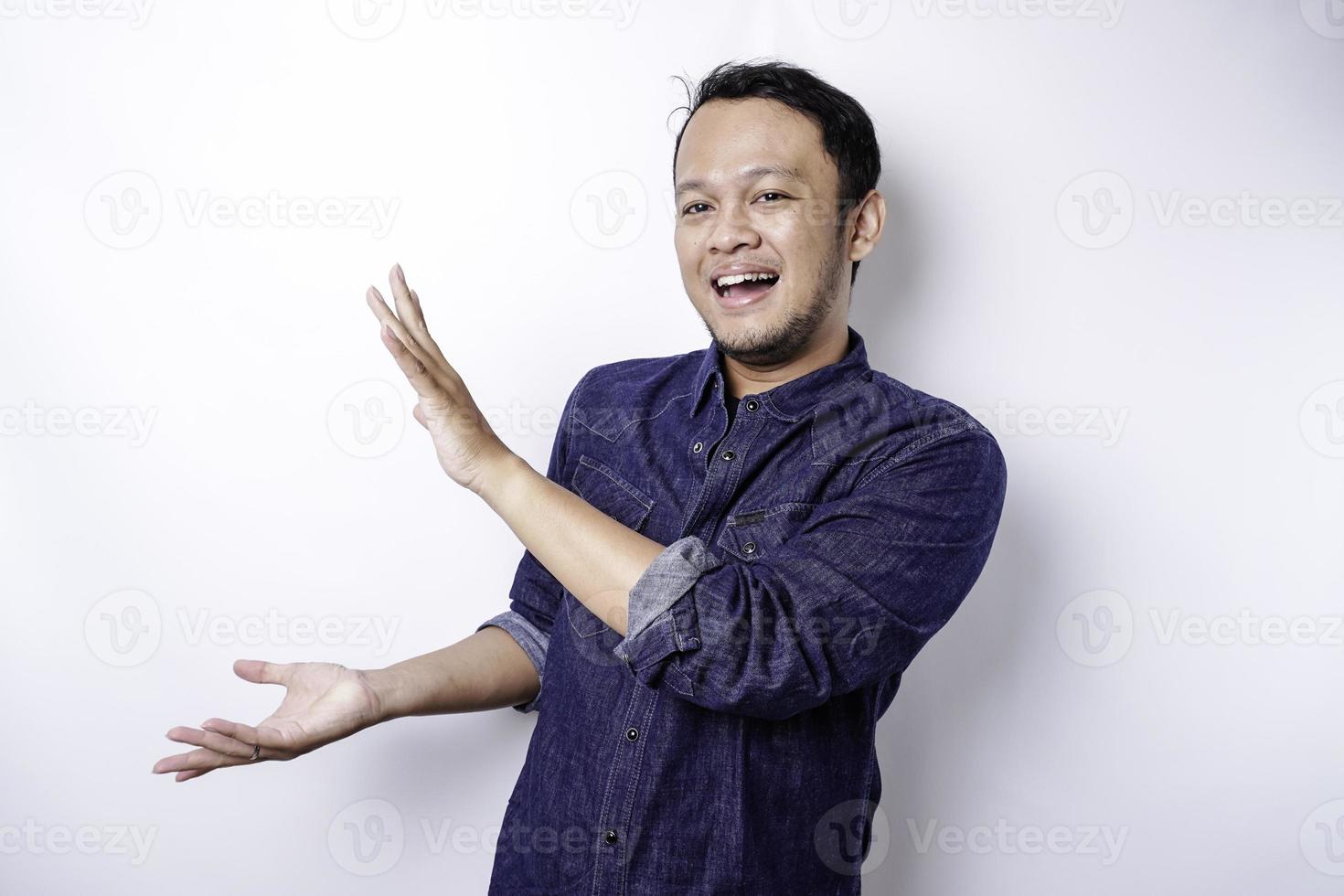 Excited Asian man wearing blue shirt pointing at the copy space beside him, isolated by white background photo