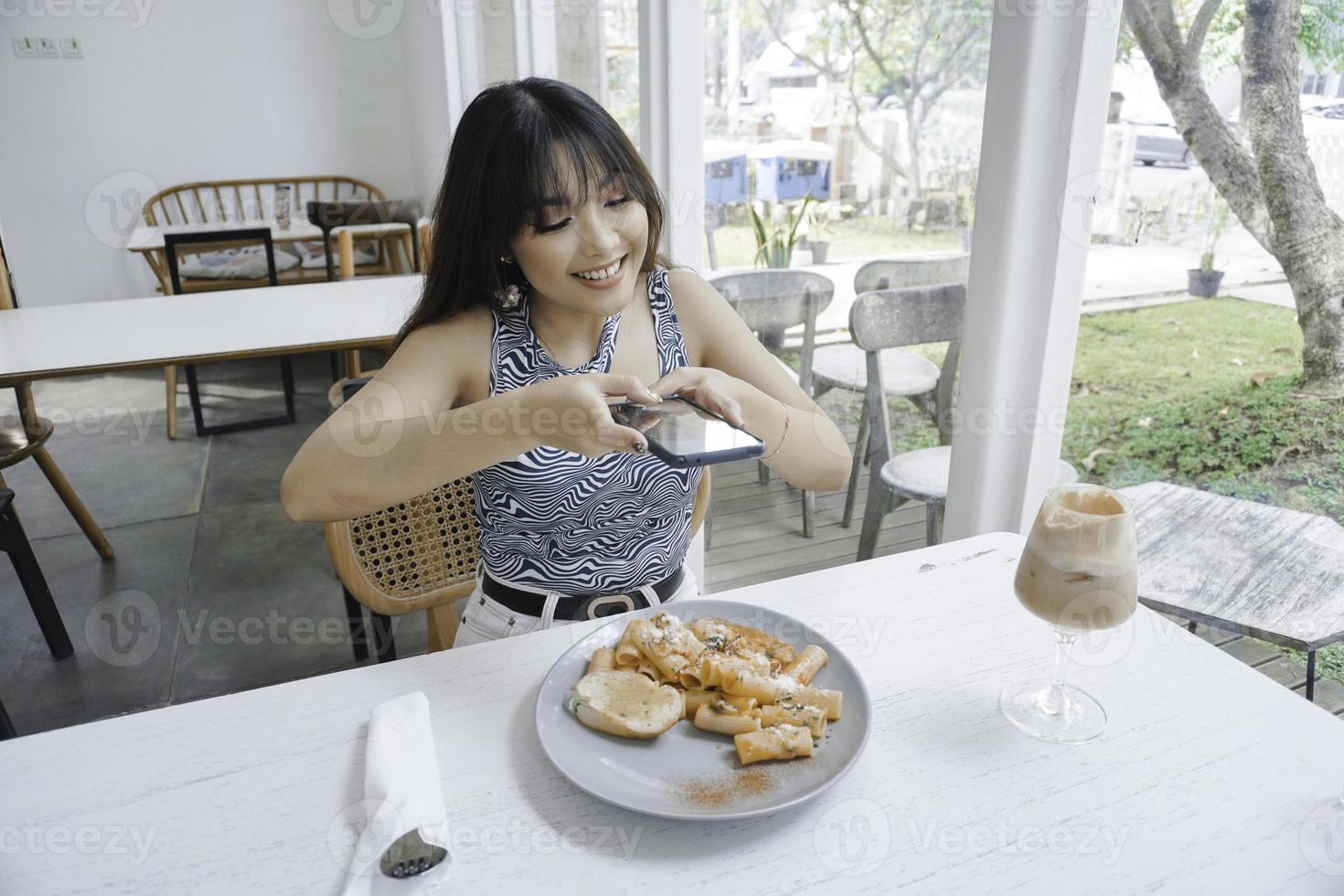 Smiling casual woman taking a picture of pasta on a white plate with mobile phone while sitting at the restaurant photo