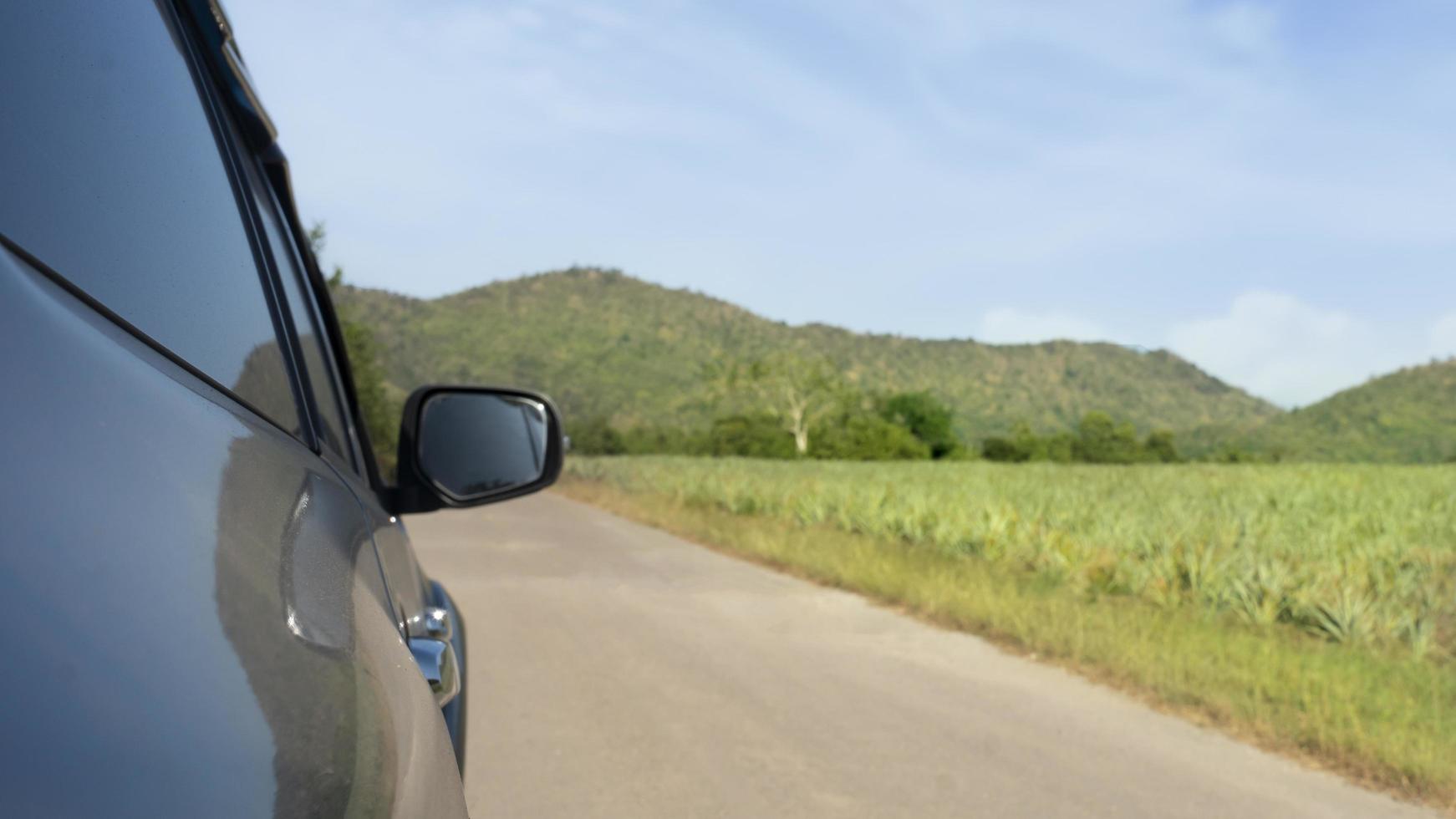 Abstract beside surface of gray car can view mirror wing of car driveing on the asphalt road. beside with blurred of pineapple plantation and distant mountains under blue sky. photo