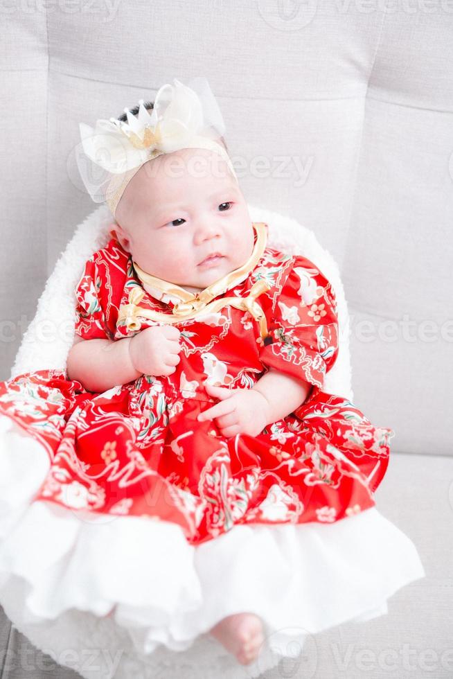 Closeup cute newborn baby in red bodysuit lying down alone on bed. Adorable infant rests on white bedsheets, staring at camera looking peaceful. Infancy, healthcare and paediatrics, babyhood concept. photo