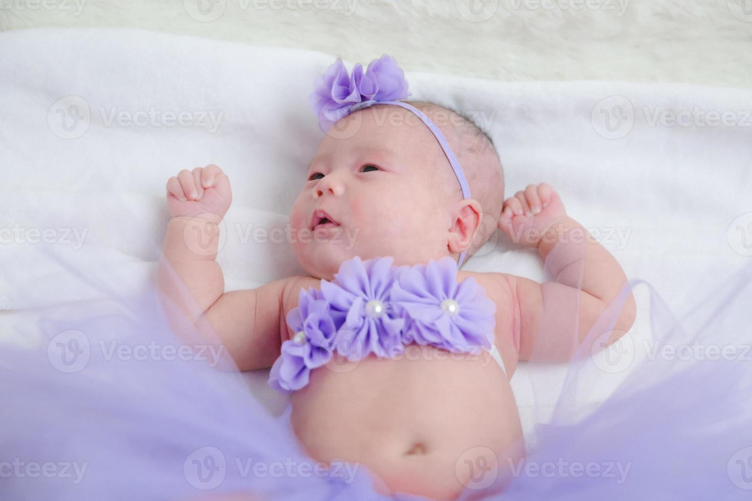 Closeup cute newborn baby in purple bodysuit lying down alone on bed.Adorable infant rests on white bedsheets, staring at camera looking peaceful. Infancy, healthcare and paediatrics, babyhood concept photo