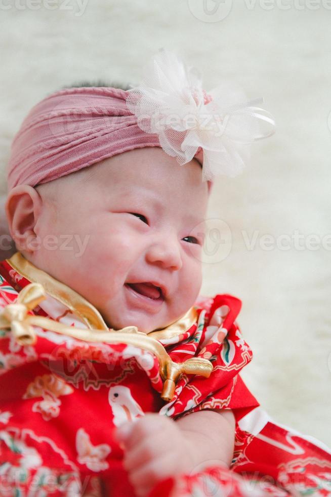 Closeup cute newborn baby in red bodysuit lying down alone on bed. Adorable infant rests on white bedsheets, staring at camera looking peaceful. Infancy, healthcare and paediatrics, babyhood concept. photo