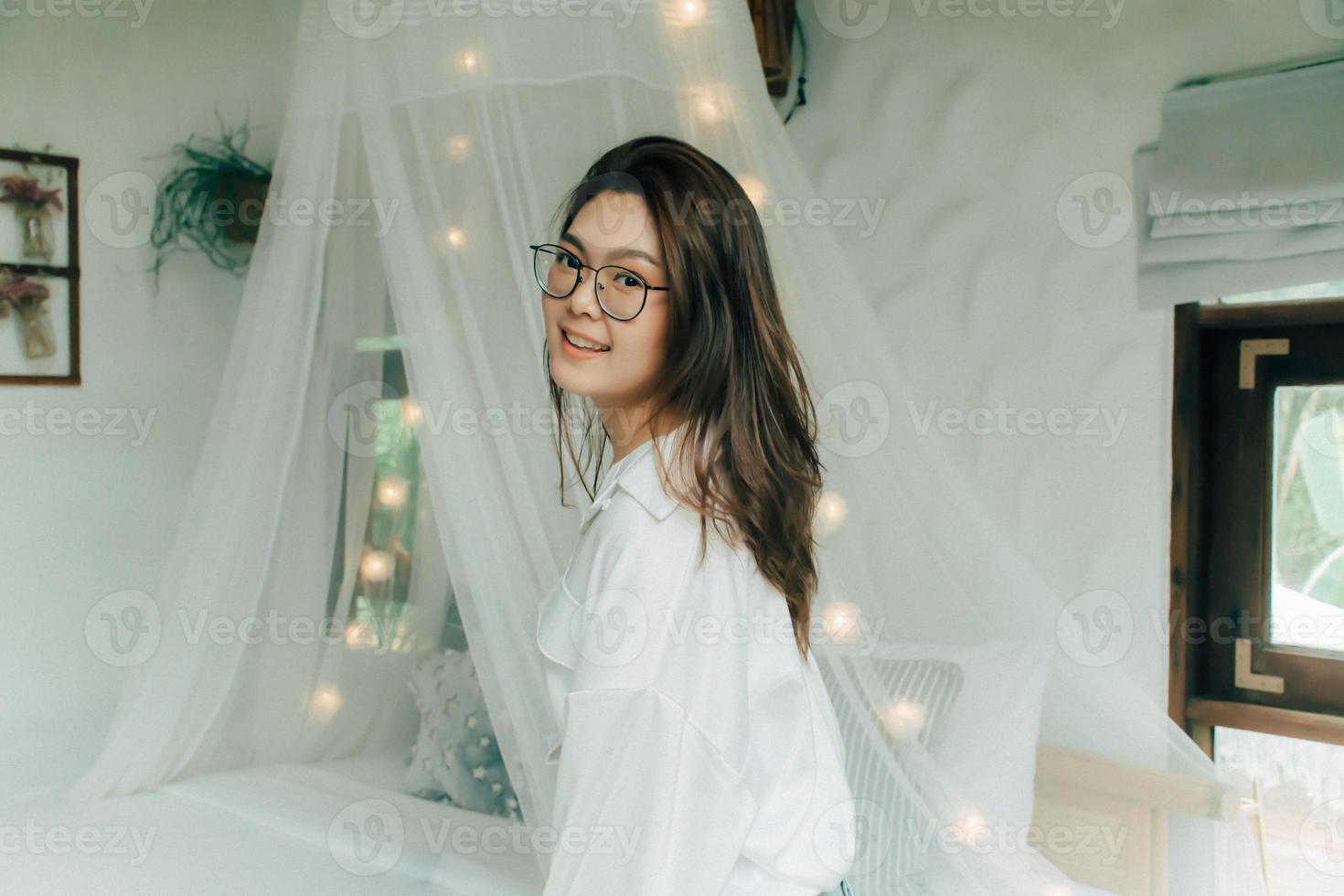 Close up portrait of woman turn around looking at camera smiles. Beautiful girl with long brown hair wearing glasses, white shirt stand beside the bed in bedroom at home. People lifestyle concept. photo