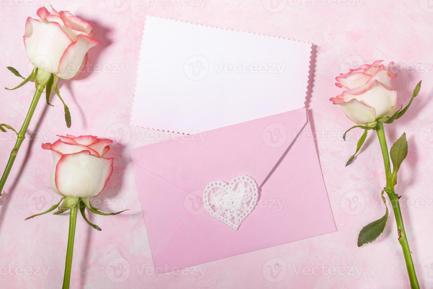 Pink envelope with empty white blank sheet and pale pink roses on pink backdrop. Copy space Top view photo