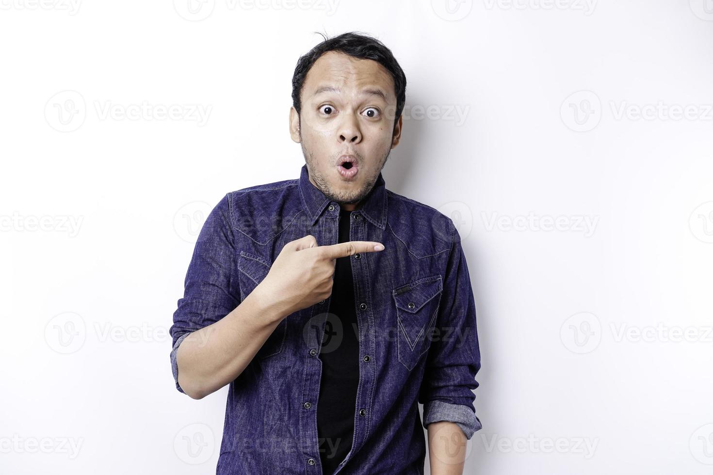 hombre asiático conmocionado con camisa azul apuntando al espacio de copia a su lado, aislado por fondo blanco foto