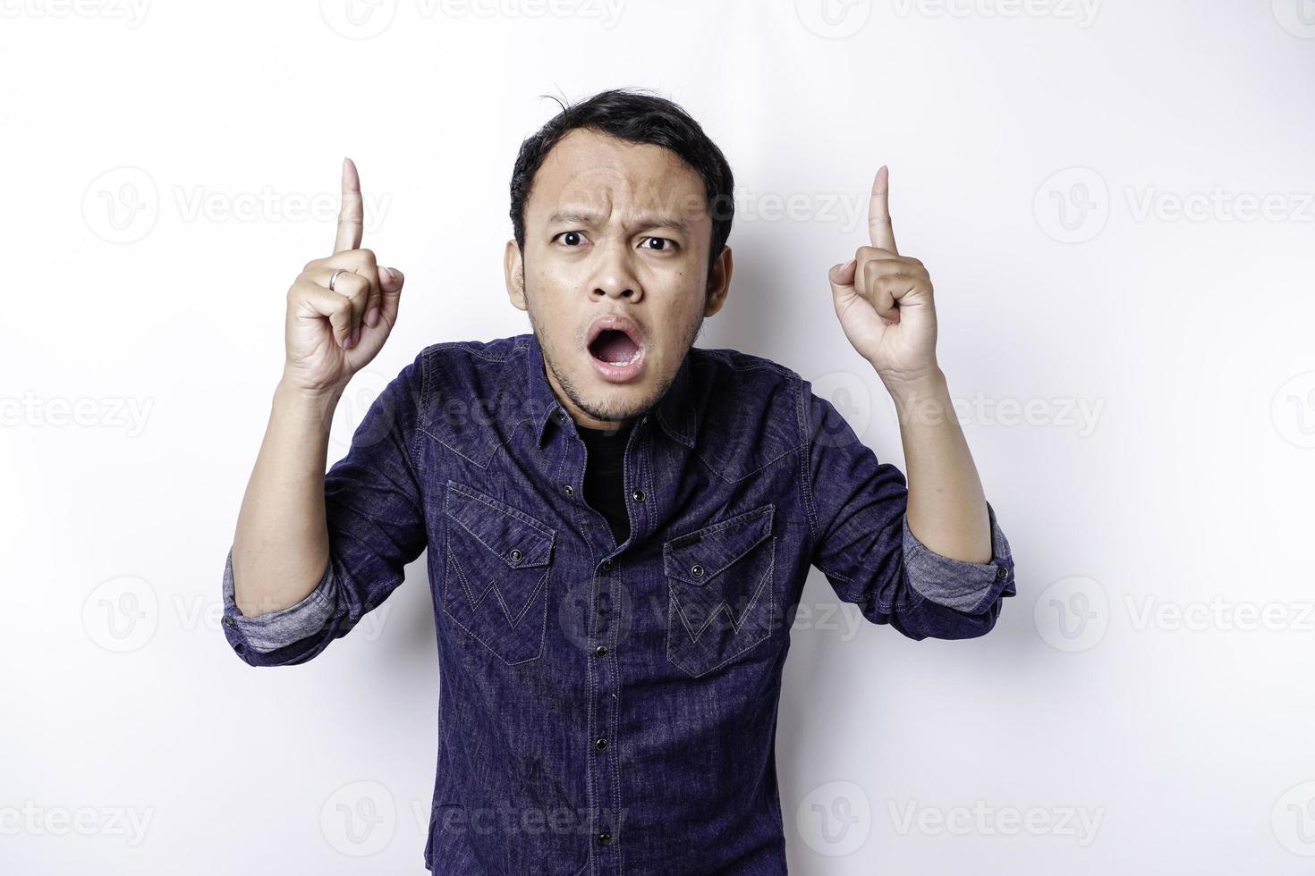 Shocked Asian man wearing blue shirt pointing at the copy space on top of him, isolated by white background photo