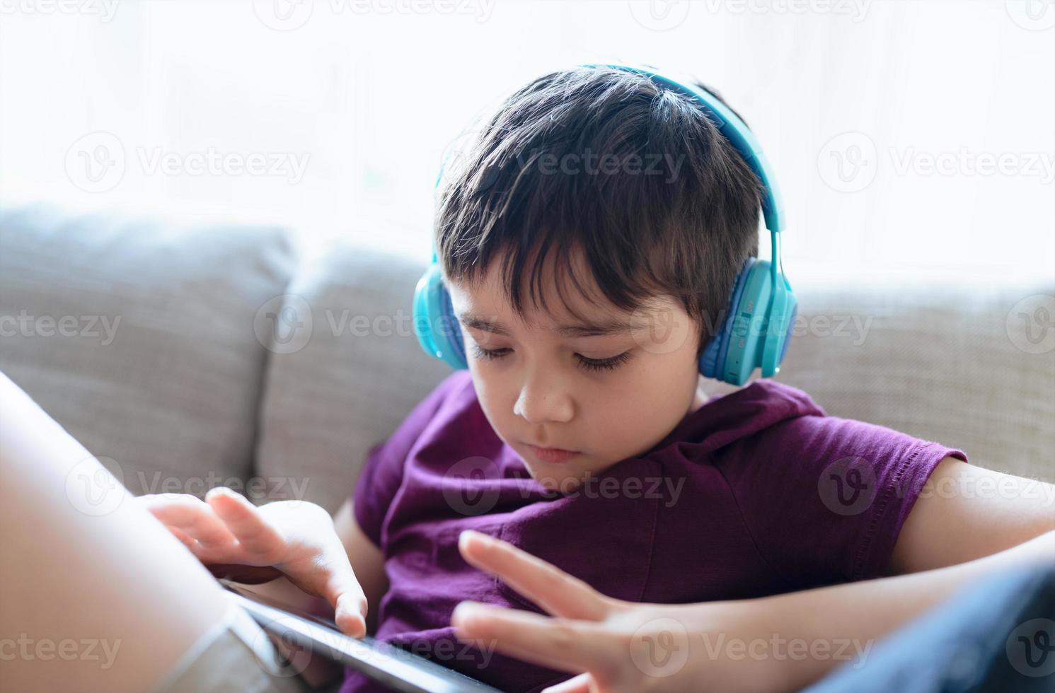 concepto de educación, retrato clave de un niño de escuela primaria leyendo un libro electrónico en una tableta para la tarea, un niño usando auriculares jugando en línea en Internet, un niño sentado en un sofá viendo dibujos animados en una libreta digital foto