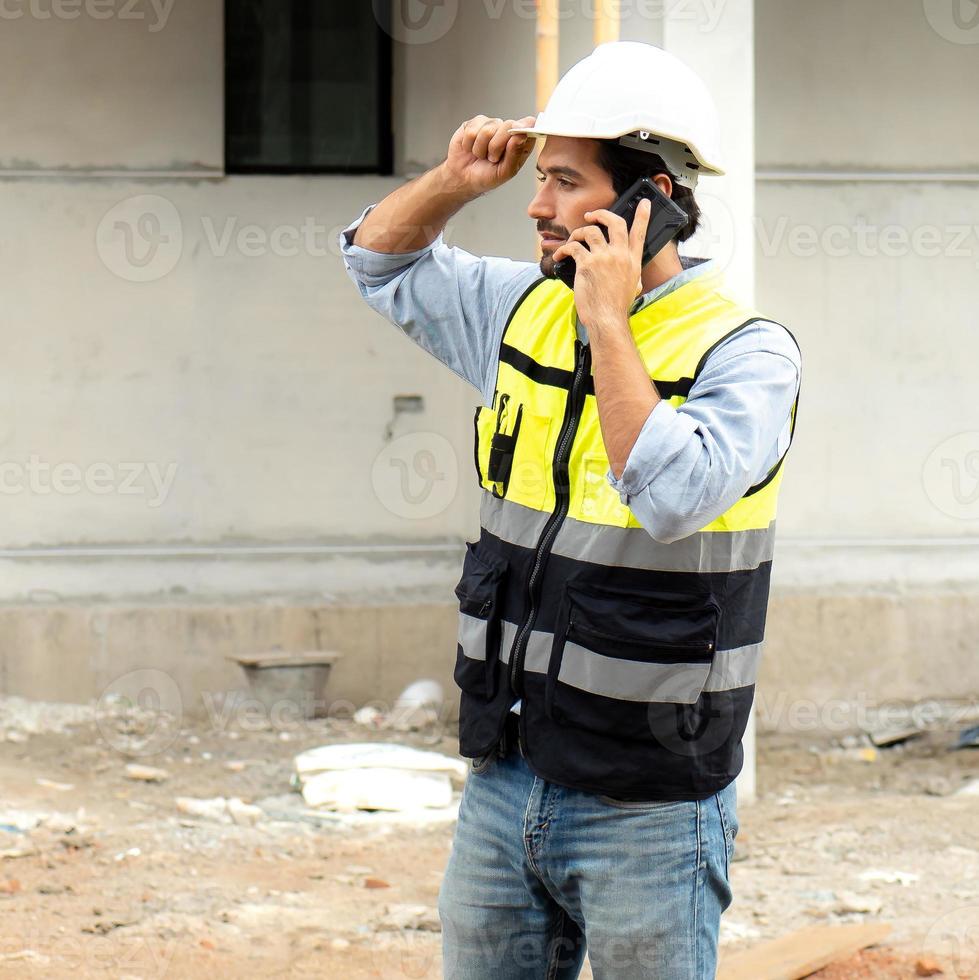 Male architect or engineer using mobile phone for communication while working at construction site. Professional building contractor standing at housing development site uses smartphone on the job. photo