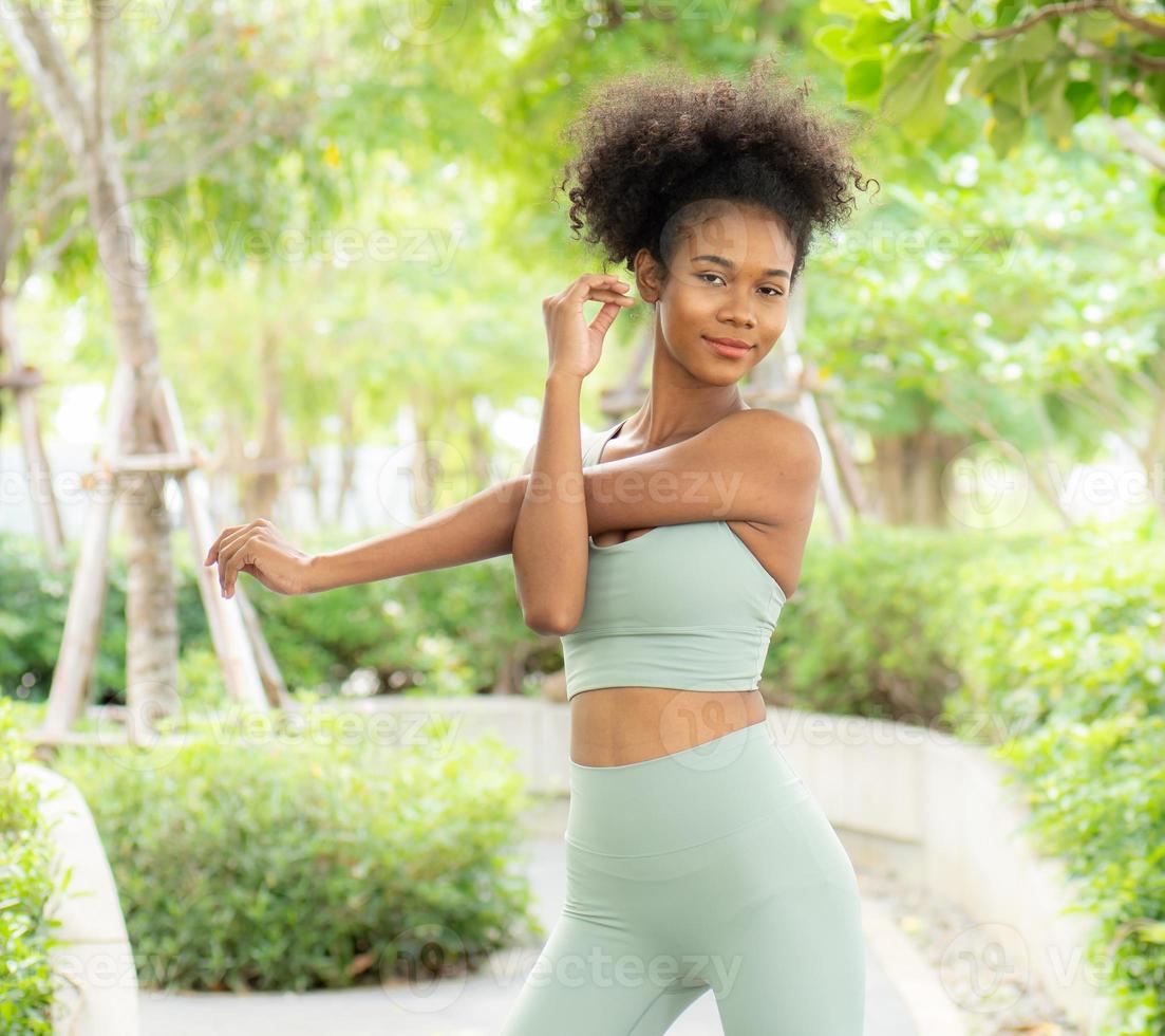 Young black woman stretching, exercising in nature park. Beautiful girl in sportswear warming by physical exercise for flexibility of muscles before jogging run in green garden. Healthy lifestyle girl photo