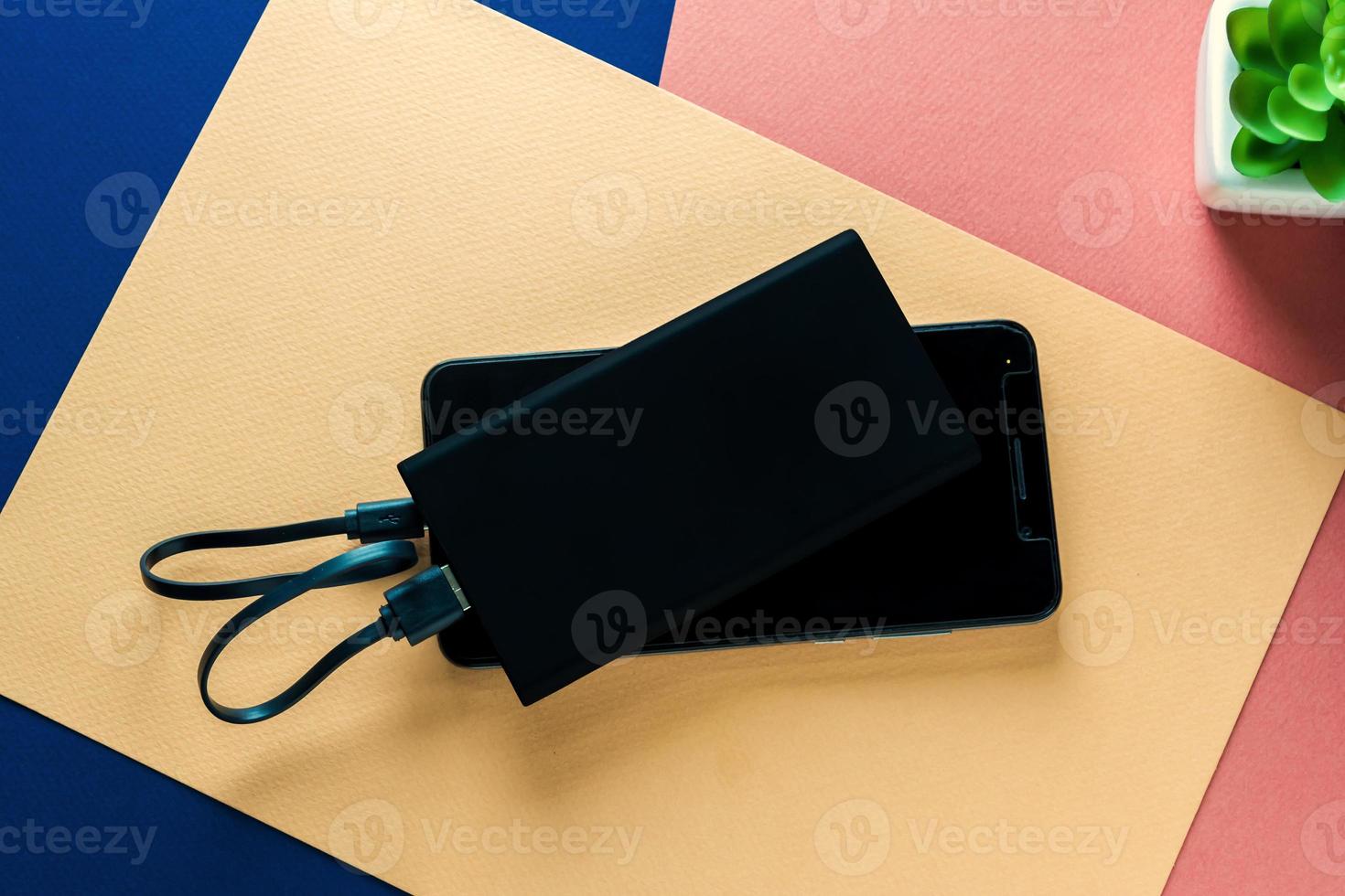 cargando un teléfono inteligente negro con un banco de energía negro sobre fondo de papel rosa, amarillo, azul. vista superior con espacio de copia, entorno de trabajo foto