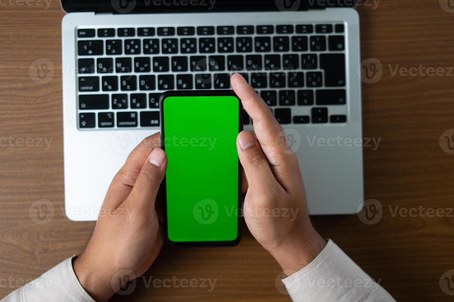 Hand holding smartphone with blank green screen  for mockups. Business desk with laptop and cell phone in flat lay photo