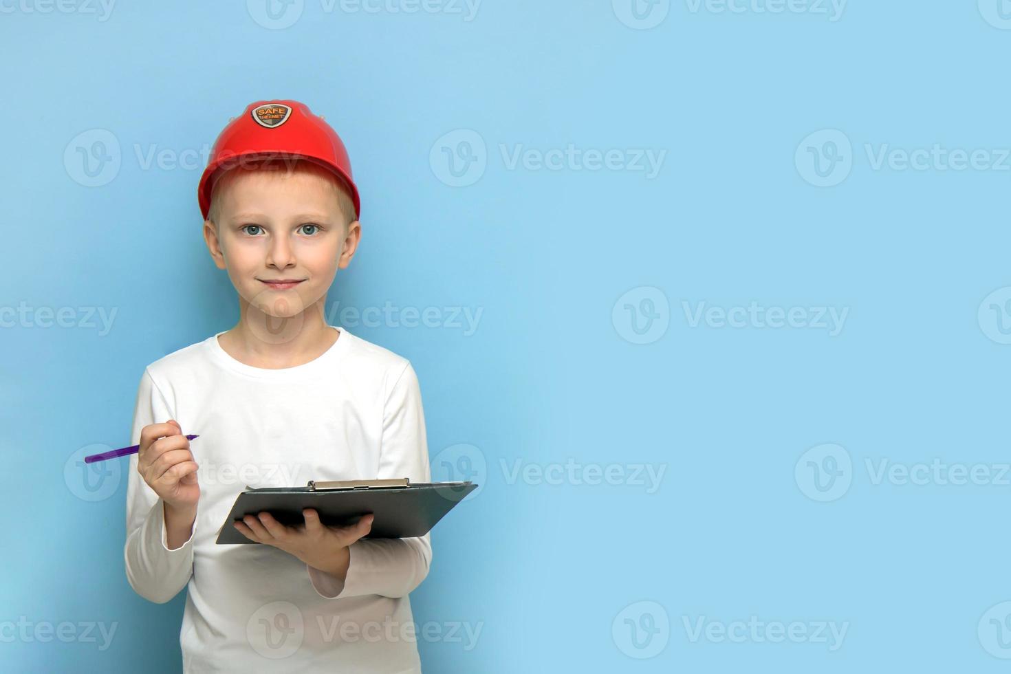 niño rubio con un casco protector de construcción con una tableta y un bolígrafo en un fondo azul con espacio de copia comprobando en un sitio de construcción foto