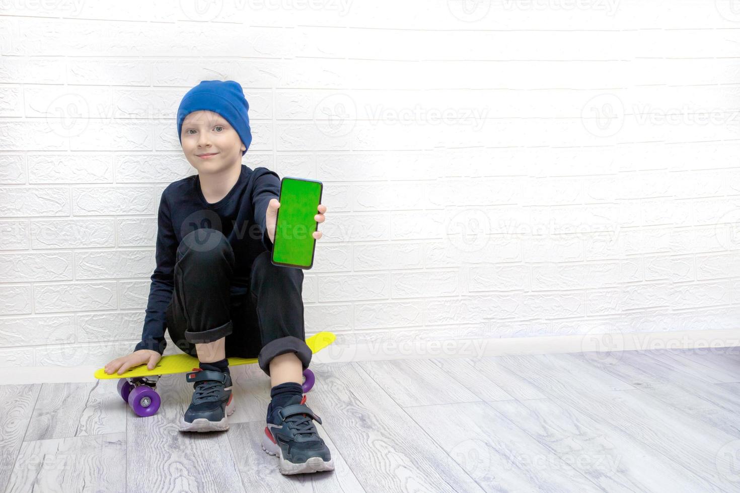 niño con un sombrero azul se sienta en una patineta sosteniendo un teléfono con una pantalla verde foto