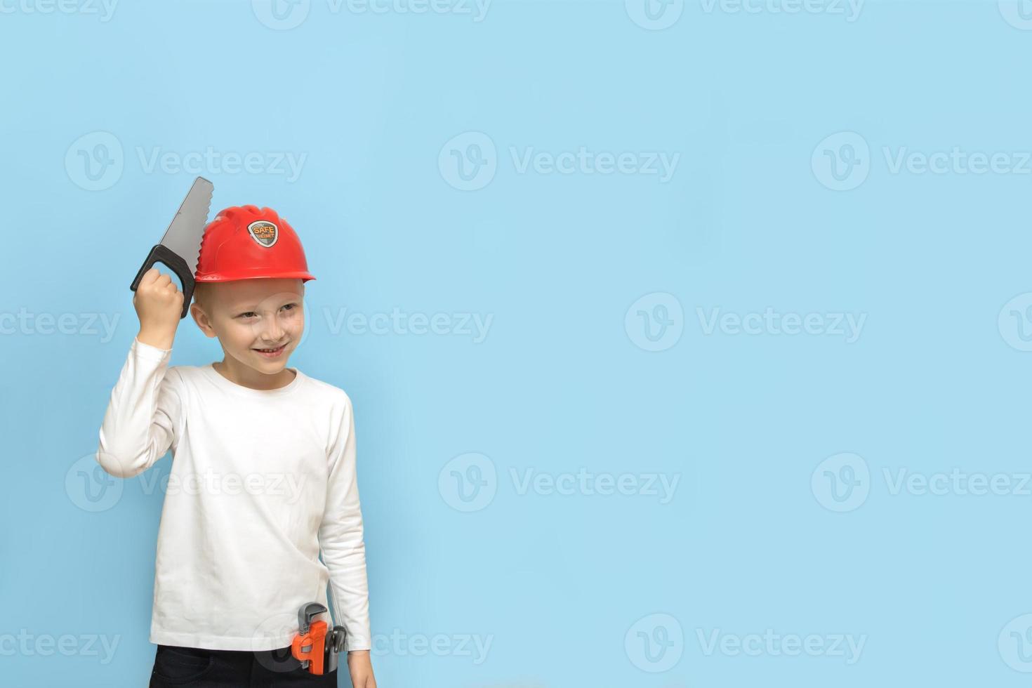 boy with a toy saw tries out a construction safety helmet on a blue background with copy space photo