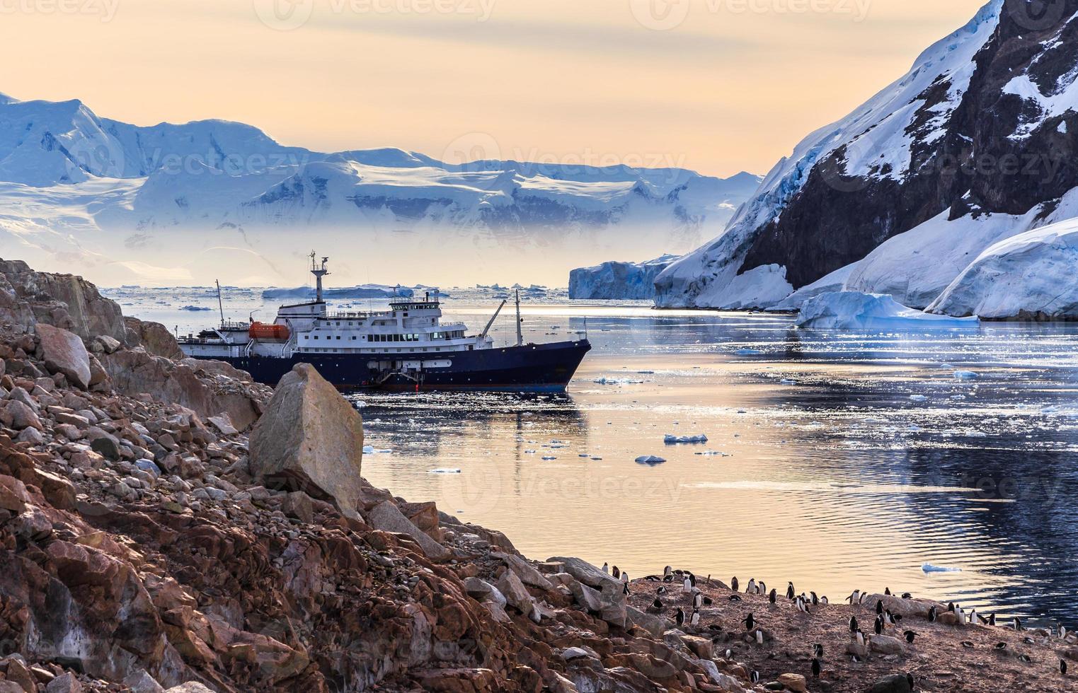 Antarctic cruise ship among icebergs and Gentoo penguins gathered on the rocky shore of Neco bay, Antarctica photo
