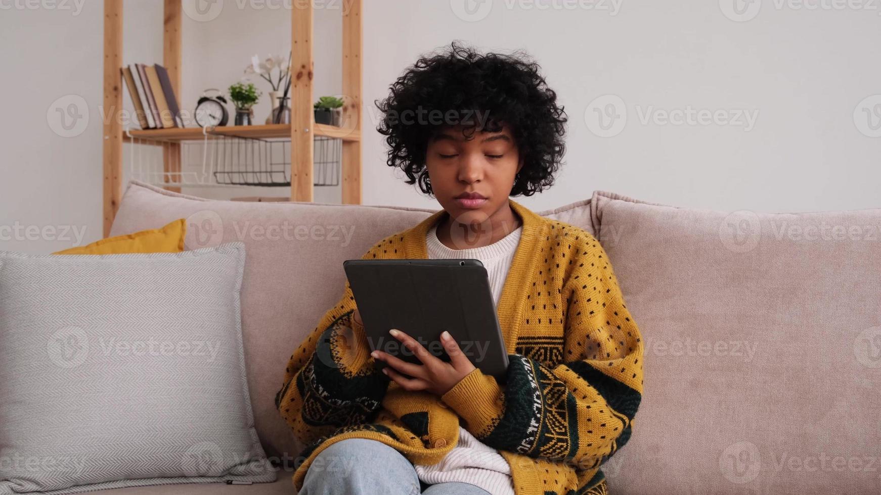 niña afroamericana sosteniendo la pantalla táctil de la tableta digital escribiendo la página de desplazamiento en casa. mujer con dispositivo de tecnología móvil navegando por Internet usando aplicaciones de redes sociales jugando. estudio trabajo compras en linea. foto