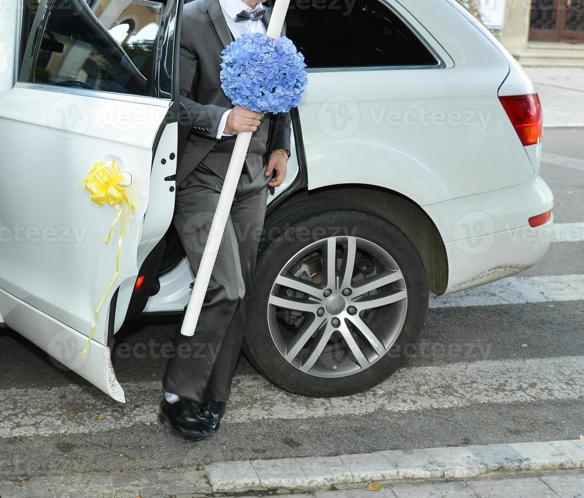 wedding groom getting out from a big white car. the man is holding a big candle with a blue flower bouquet photo